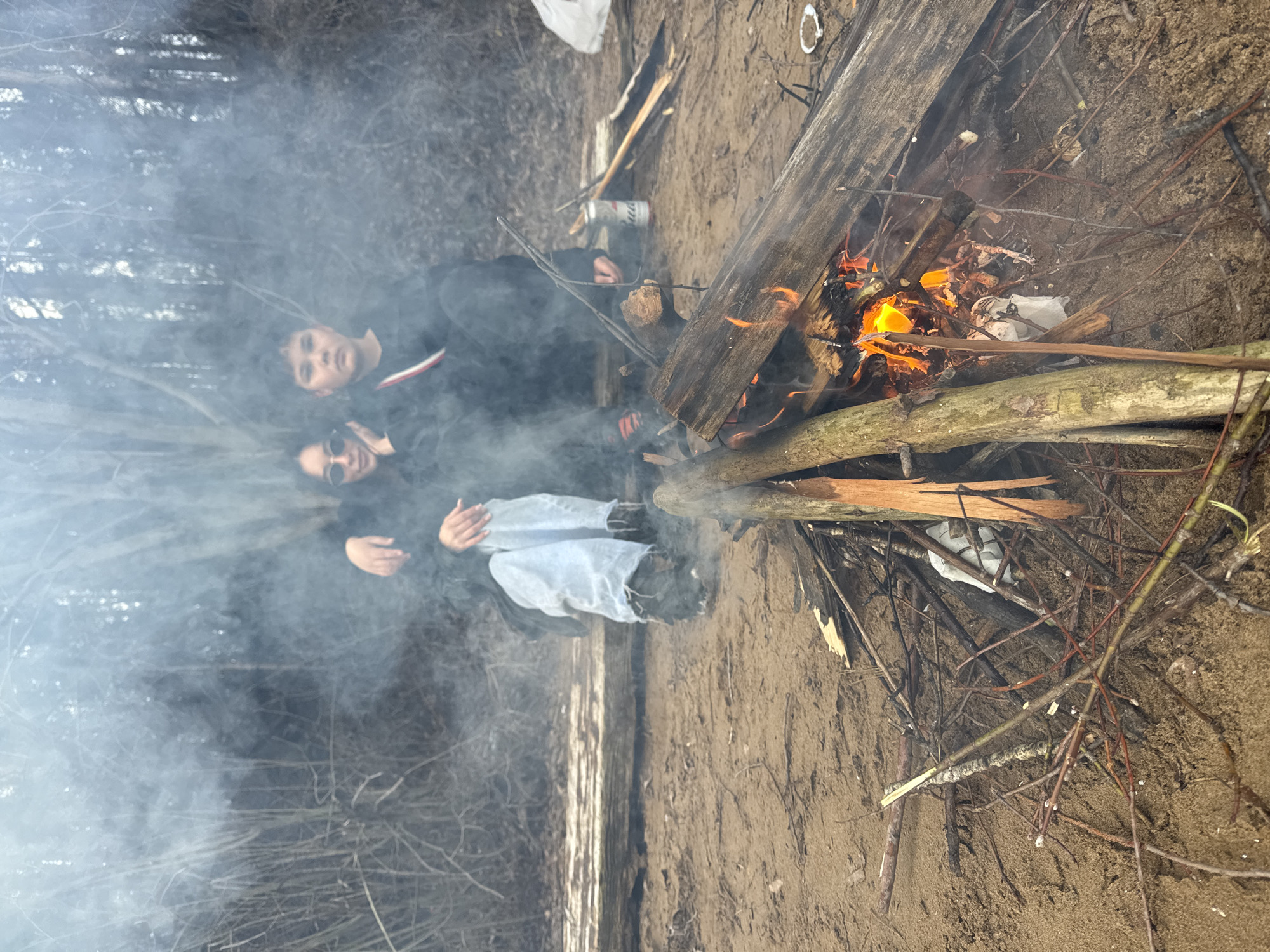 A photograph shows two individuals positioned near a campfire. The setting seems to be an outdoor area, potentially a beach or wooded area, given the sandy ground and presence of trees visible through the smoke.
One person is wearing sunglasses and a dark jacket or hoodie and appears to be gesturing with their hands. The second person is wearing a dark-colored top with a light-colored stripe on the chest. The campfire is constructed of logs and branches. The fire is burning actively. Thick smoke emanates from the fire. There are some discarded items in the background, including cans.
While this image does not show definitively this is in Russia or near the city of Chaikovsky, Russia, the trees and sand may align with this location.