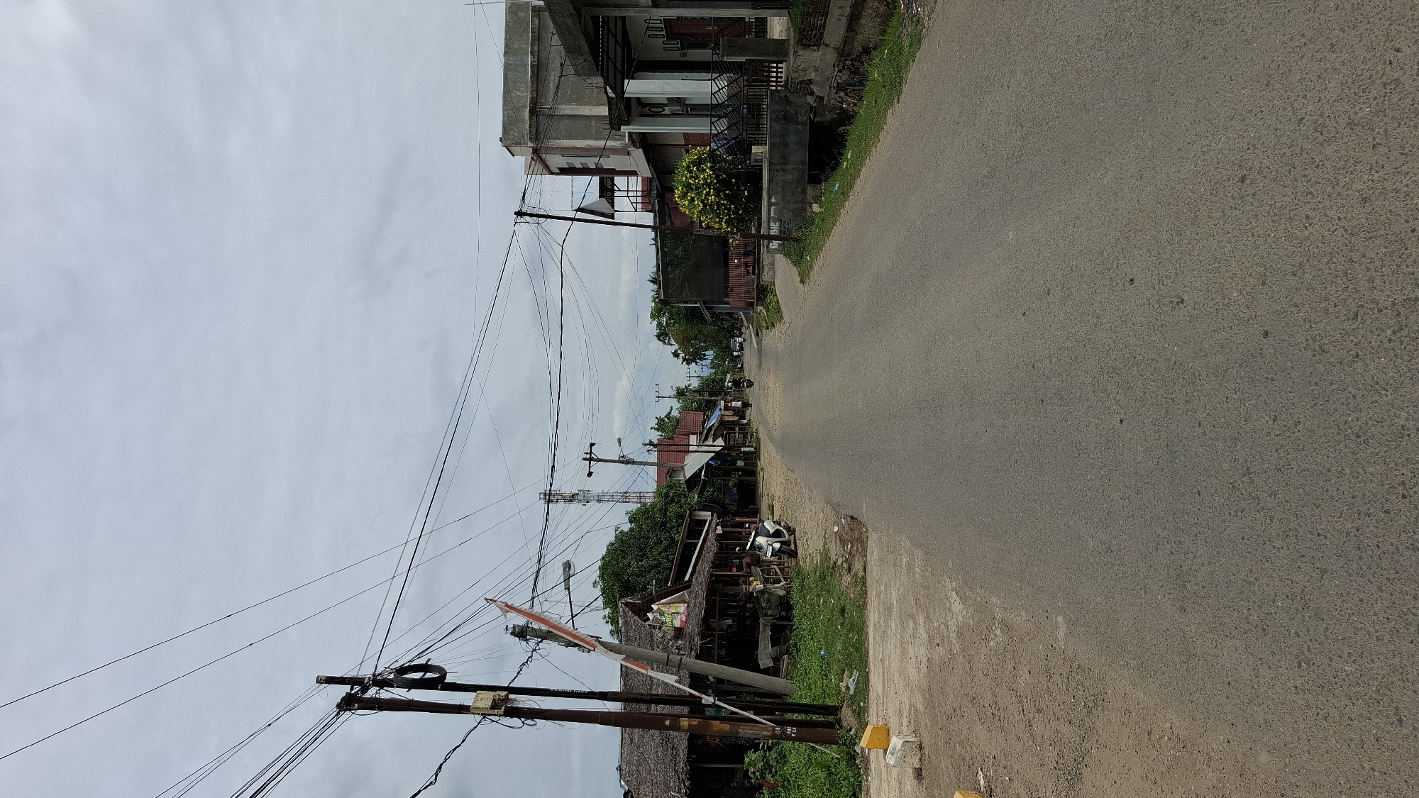 This is a medium-angle, eye-level shot of a paved road in Bale, Indonesia. The road extends into the distance, flanked by buildings and vegetation on both sides. The sky is overcast and gray. On the left side of the road, there are several small, rustic buildings with thatched roofs, and a white motorcycle is parked in front of one of them. A utility pole with numerous wires is also visible on the left. On the right side of the road, there are more modern buildings, including a two-story structure with a concrete facade. A few people are visible in the distance, one riding a motorcycle. The overall impression is a rural or semi-rural street scene.
