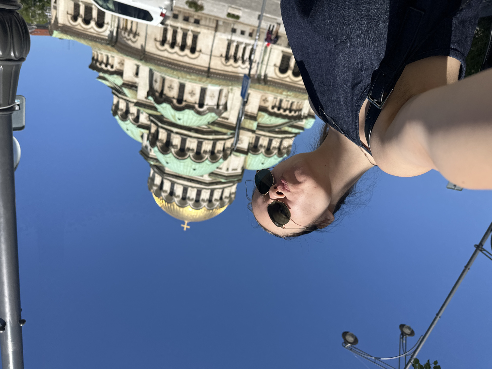A low-angle selfie shot shows a woman with dark hair pulled back wearing sunglasses, with a cathedral in the background against a clear blue sky. The cathedral is the Alexander Nevsky Cathedral in Sofia, Bulgaria. The woman wears a dark blue top with suspender-like straps. A white van is partially visible in the upper portion of the background. In the lower right, there is a streetlight with two lights. A lamp post is on the left edge of the image.