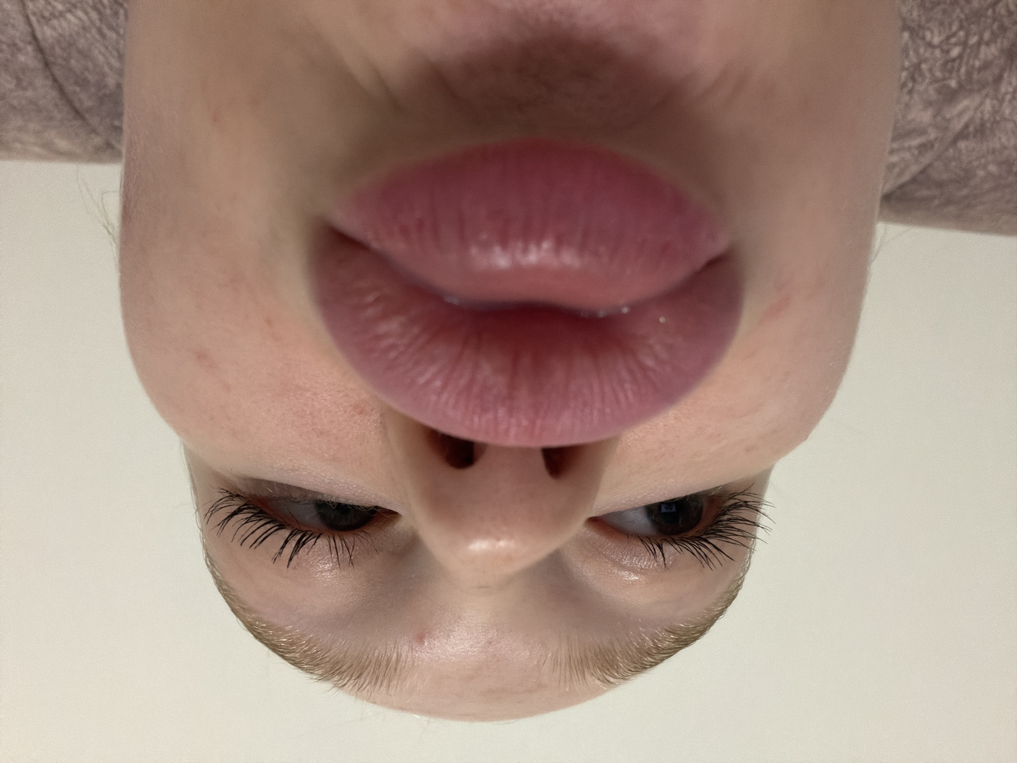 A close-up, inverted perspective of a person's face, dominated by the lips and nose. The lips are full and appear to be coated with a lip product. The eyes are visible below the nose, with dark eyelashes. The skin is light-toned and shows some texture. The location is not discernible.