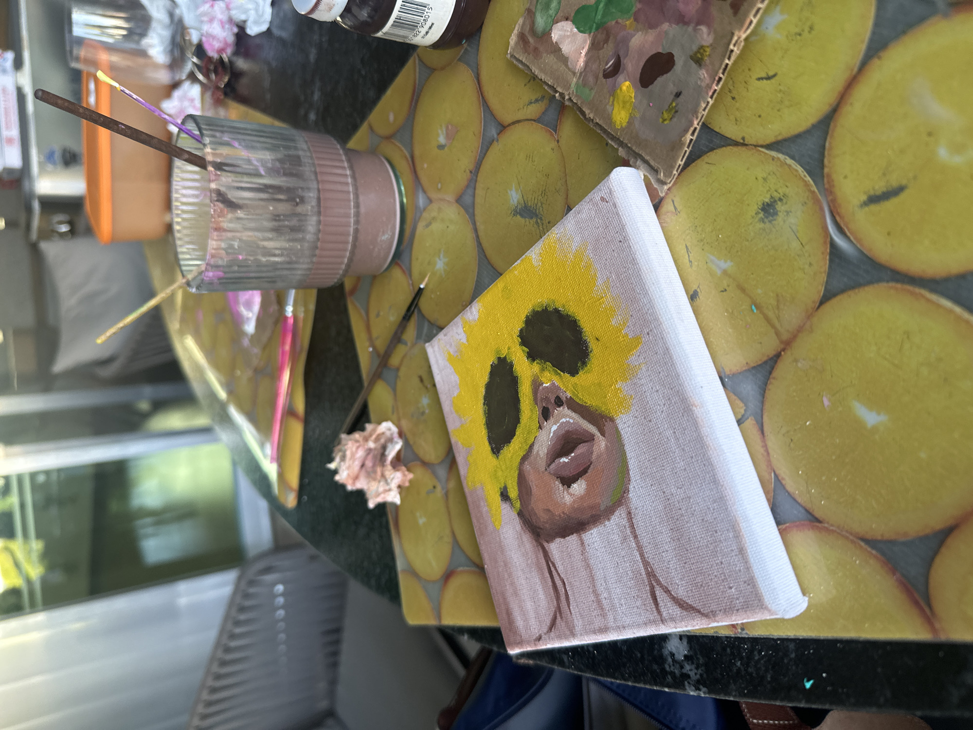 The image depicts a close-up of an art project in progress, set on a dark, speckled countertop. In the foreground, a small, square canvas displays a partially painted sunflower with a brown center. Next to it, another smaller canvas or palette shows dabs of various paint colors. A glass jar filled with a brownish liquid holds several paintbrushes, some with paint residue on them. A small wad of pinkish material, possibly a used paper towel or cloth, rests on the countertop near the jar. Behind these items, a yellow mat with circular patterns is visible. In the background, a blurred scene shows outdoor elements, including what appears to be a window or glass door with reflections of greenery and possibly a building. The overall setting suggests an art studio or a creative space, potentially outdoors given the background. The presence of art supplies like paint, brushes, and canvases indicates an active art-making process. The location is identified as San Bernardino Tlaxcalancingo, Mexico.
