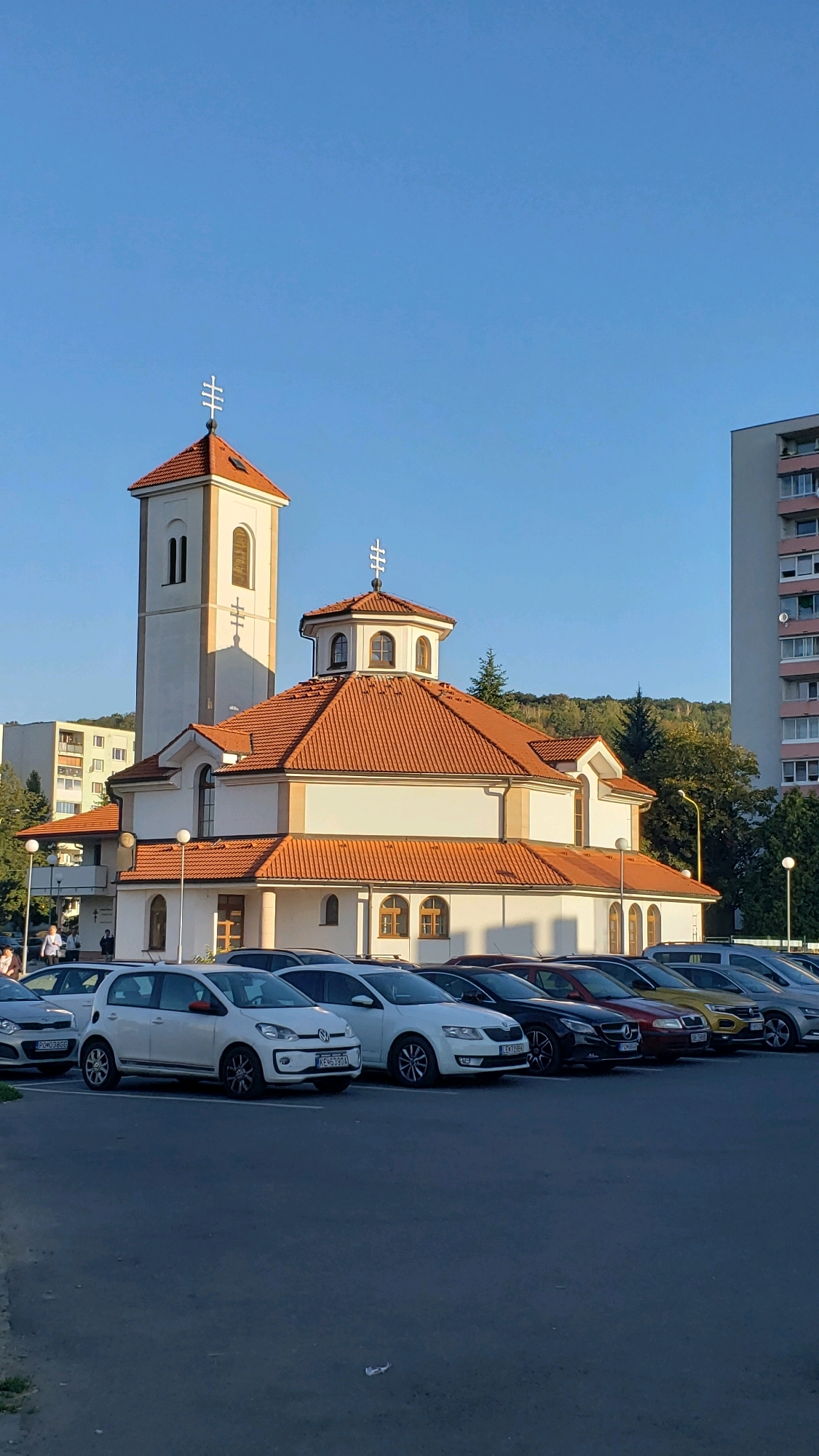 The image depicts the Church of Saint Nicholas in Prešov, Slovakia, situated in a built-up urban environment. The church, with its prominent bell tower and terracotta-tiled roof, is the central subject. A line of parked cars is visible in the foreground, with some buildings and a wooded hill in the background. The sky is clear and blue, suggesting it is daytime, likely late afternoon given the warm sunlight casting shadows. A few individuals are seen near the entrance of the church. The scene is static and appears to capture a typical day in this location.