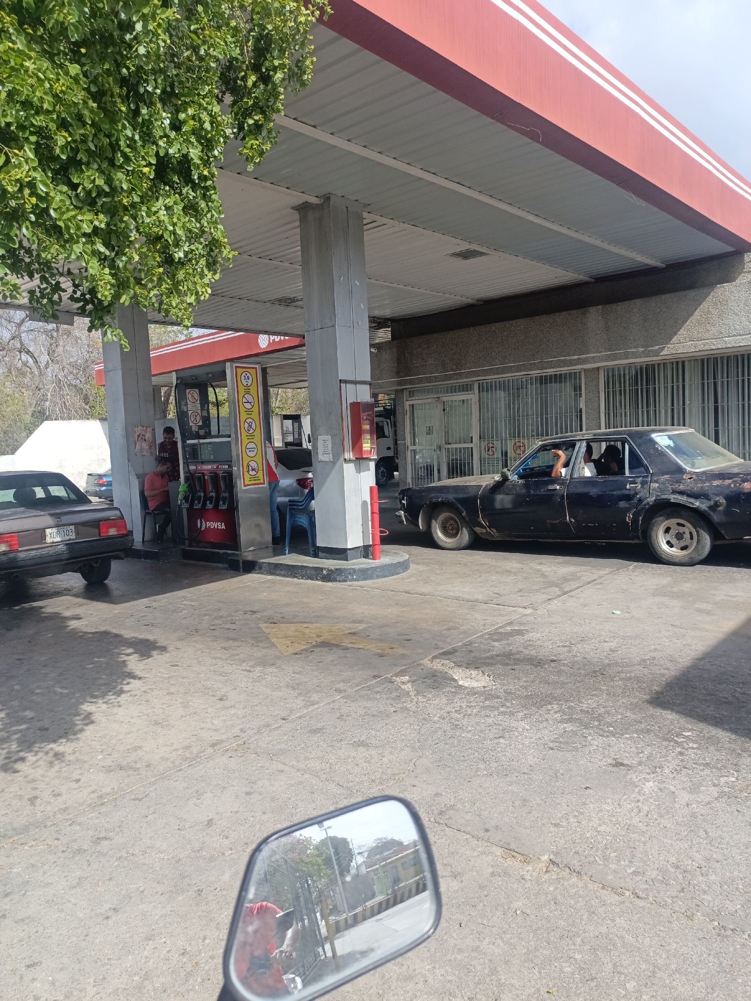 The image depicts a gas station in La Tagua, Venezuela. A dark-colored sedan is parked near a building with barred windows, while a brown car is parked at a PDVSA pump. Two individuals are visible near the pump, one seated. The gas station has a red and white roof. A motorcycle's side mirror is visible in the foreground. The scene appears to be outdoors with sunlight.