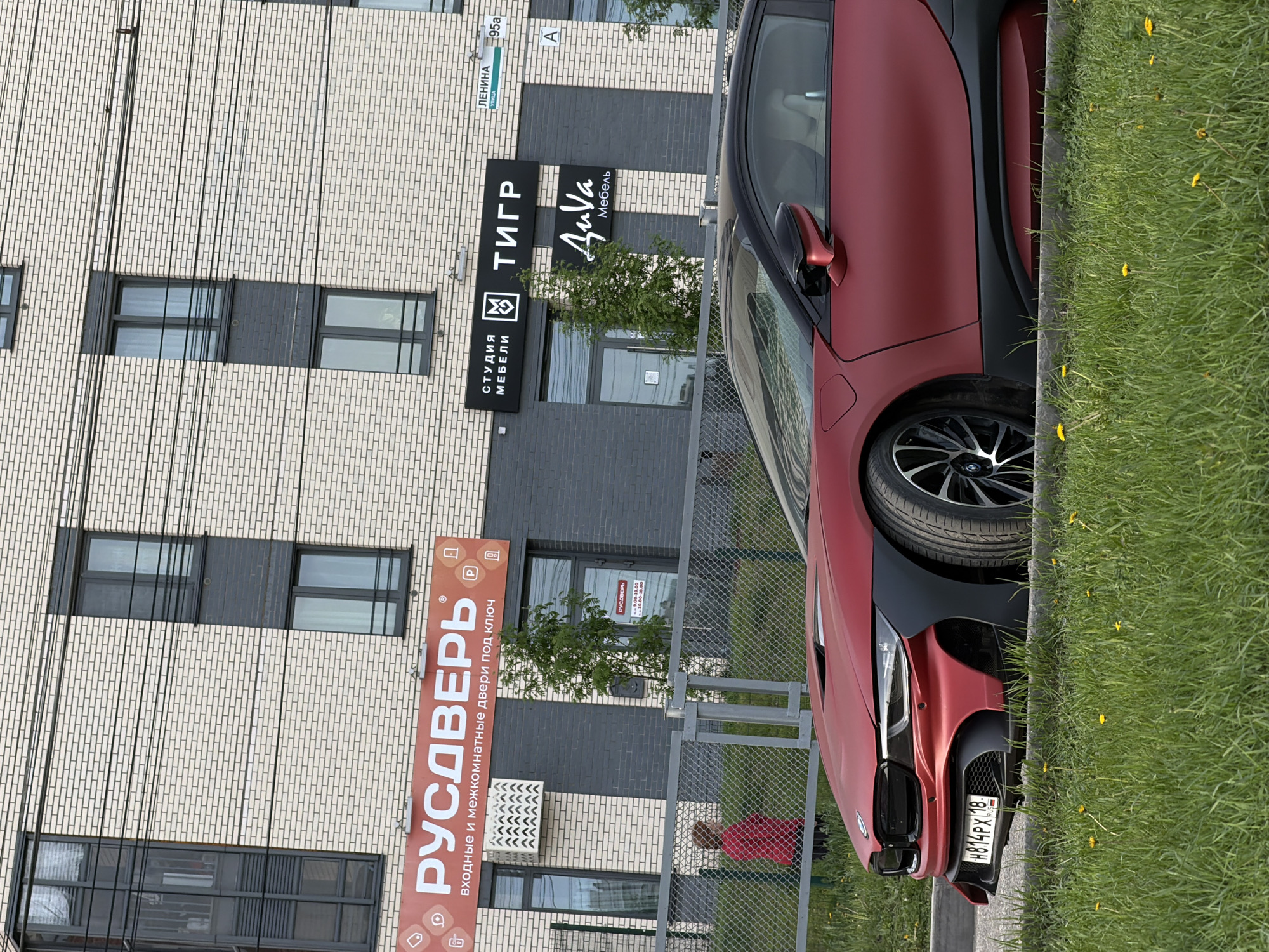 The image shows the side of a red BMW i8 parked next to a patch of grass. In the background is a building with multiple businesses. There are signs for "PyCDBEPb" (doors), "Студия Мебели" (Furniture Studio), "ТИГР" (Tiger), and "ДиѴa Мебель" (DiVa Furniture). The address is "ЛЕНИНА 95a" (Lenin Street 95a). The license plate on the car reads "H814PX 18" with a Russian flag. There is a person in a red shirt standing behind a fence in front of the building. The scene is located in Izhevsk, Russia based on the license plate and street signs.