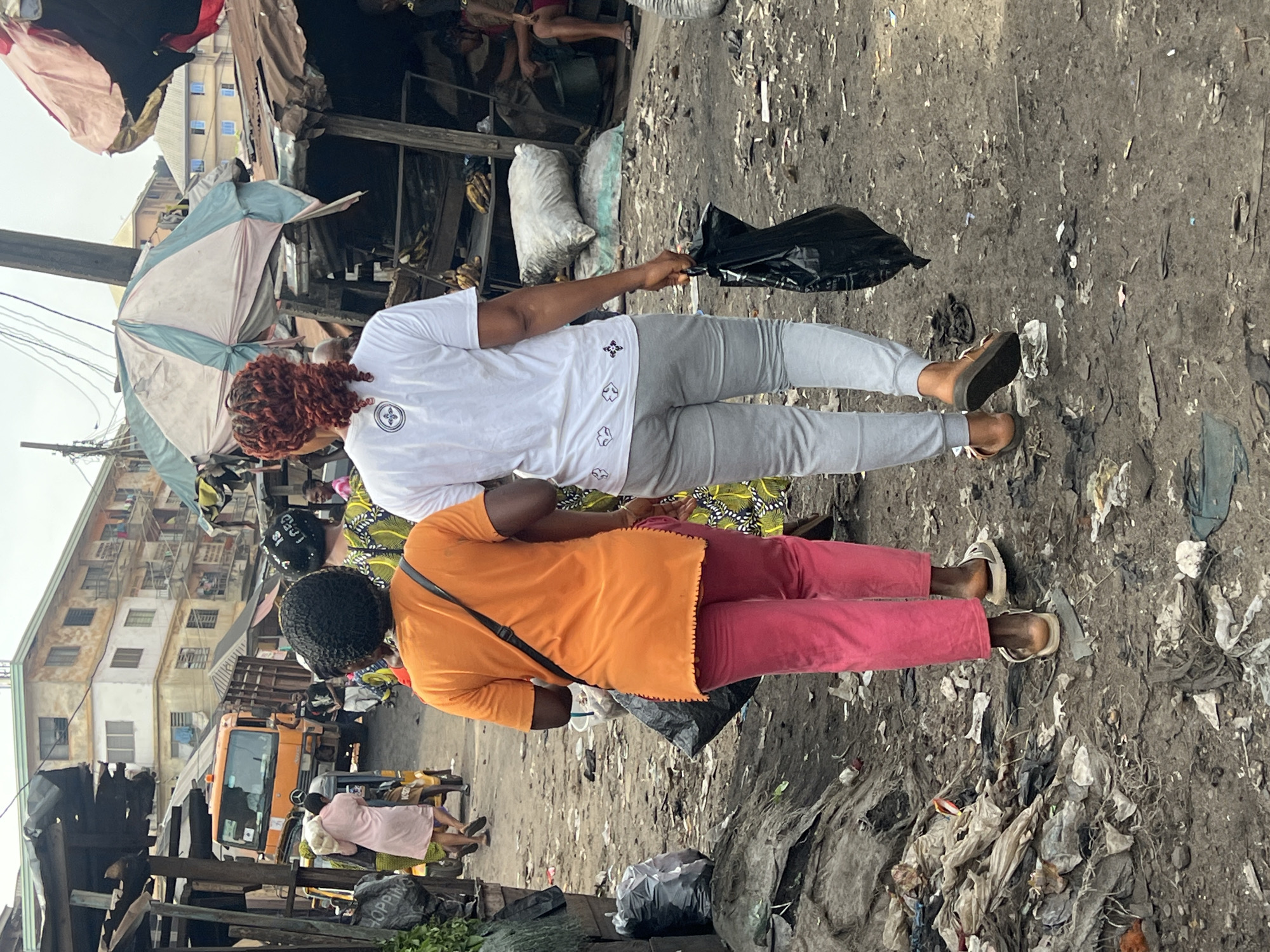 The image shows two women standing on a litter-strewn ground, possibly a market area. One woman wears a white t-shirt and grey pants, holding a black plastic bag. The other woman wears an orange top and pink pants, with a strap across her shoulder. In the background, there are stalls, buildings, and an orange truck. The location appears to be a crowded, urban environment with signs of poverty and poor waste management.