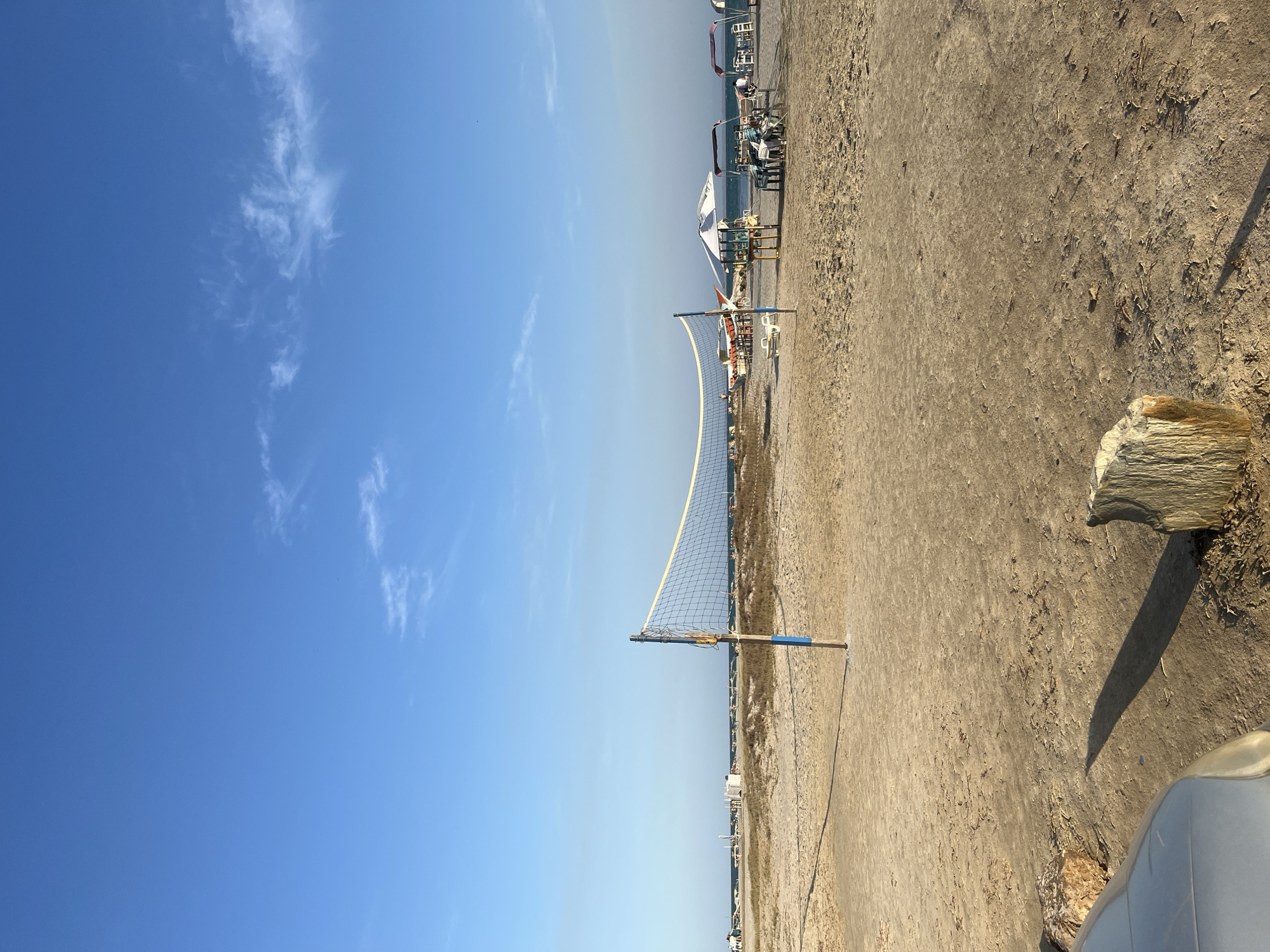 The image depicts a sunny day at a beach in El Yaque, Venezuela. The sky is clear blue with scattered white clouds. A volleyball net stands on the beach, dividing the scene. Behind the net are beach umbrellas and chairs, suggesting a recreational area. In the foreground, the beach is sandy and textured, with some debris present. A piece of driftwood lies to the right. Part of a car is visible in the very bottom right corner.