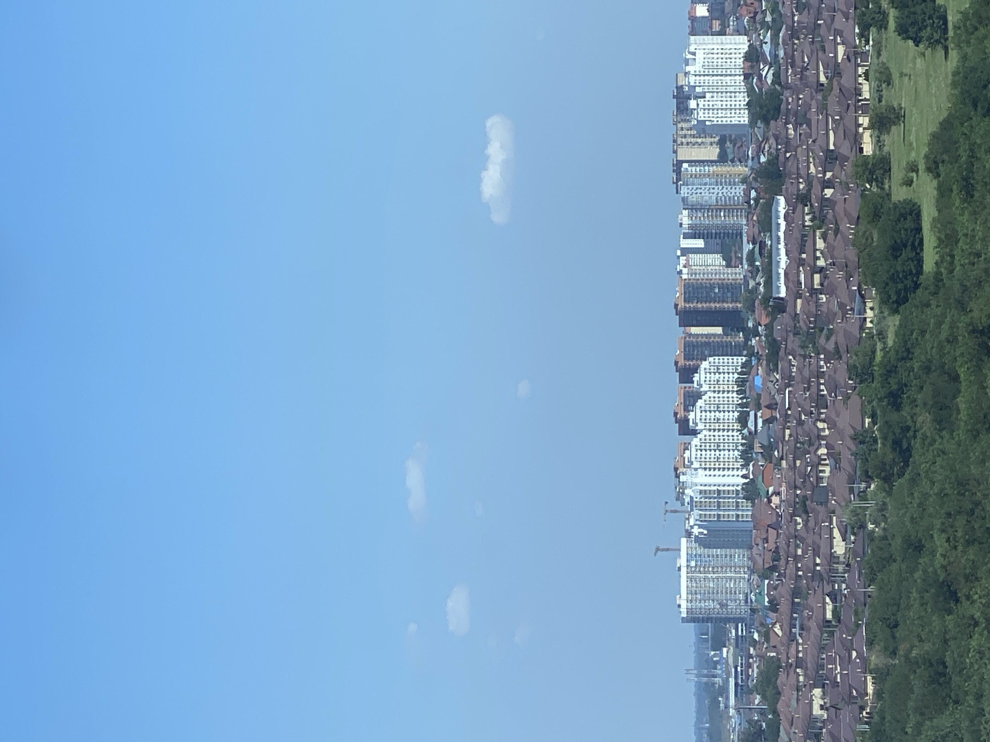 An eye-level, medium shot captures a sprawling suburban landscape beneath a clear blue sky with a few scattered white clouds. In the foreground, a dense, verdant forest covers the lower portion of the frame. Behind the trees, a vast expanse of closely packed, single-family homes with consistent reddish-brown tiled roofs dominates the midground. These houses are uniformly beige or light yellow, creating a monotonous, repeating pattern. Beyond the residential area, a cluster of taller, modern apartment buildings rises in the background, their facades displaying a mix of white, beige, and reddish-brown hues. Construction cranes are visible amongst the apartment buildings, suggesting ongoing development. The overall impression is one of a rapidly expanding urban area characterized by low-density housing and modern high-rise construction.