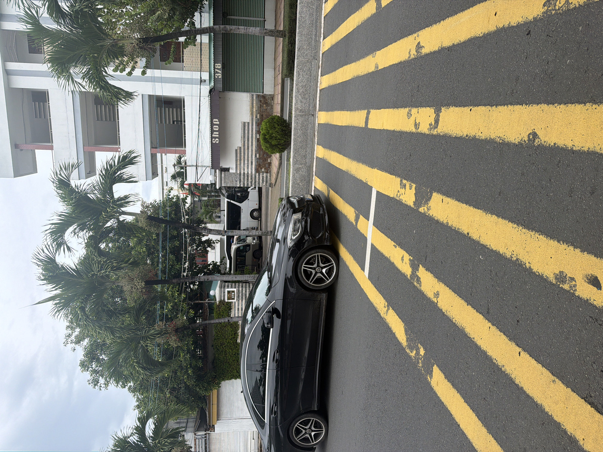 The image captures an urban street scene, likely in Nha Trang Ward, Vietnam, on an overcast day. A black car, possibly a Mercedes-Benz given the wheel emblems, is parked on the side of a paved road marked with bold, faded yellow stripes. A single white line is also visible on the road surface near the car.
To the left of the parked car, a grey concrete curb separates the road from a sidewalk area. Further back, a multi-story white building with multiple rectangular openings or balconies dominates the background. On the ground level of this building, an awning displays the word "SHOP" written vertically, and the number "378" is visible on the building's facade. A small, neatly trimmed round bush sits on a patch of greenery in front of the building.
Numerous lush green palm trees and other dense foliage are scattered throughout the scene, suggesting a tropical or subtropical climate. Power lines are visible crisscrossing the upper part of the image. In the background, partially obscured by trees and other elements, a white van is also parked. The overall impression is a quiet, stationary daytime street view with no discernible human activity.