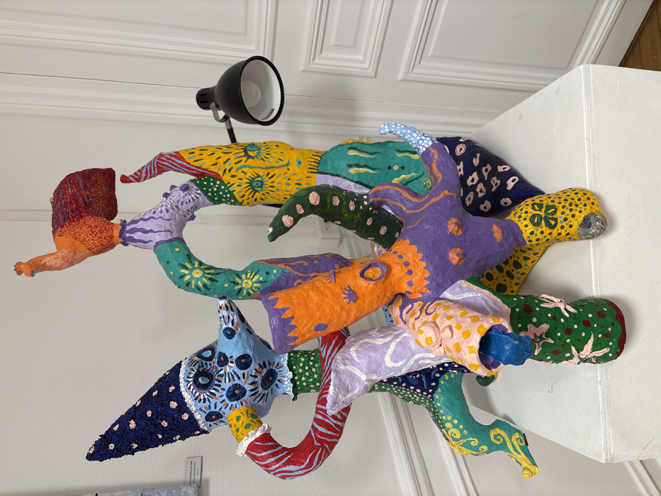 An intricately detailed, multi-component sculpture is displayed on a white rectangular pedestal within an interior setting in Villejuif, France. The sculpture, likely constructed from papier-mâché or a similar textured material, presents a complex assemblage of intertwined, stylized organic and anthropomorphic forms. It is rendered in a vibrant, extensive palette of colors including blues, reds, greens, yellows, oranges, purples, and blacks, often adorned with dots, stripes, and intricate patterns. Prominent features include a blue-faced figure on the left with multiple circular 'eyes' and a dark blue conical hat dotted with pink, an orange-red bird-like form situated at the sculpture's apex, and numerous interconnected limbs or appendages exhibiting diverse patterns. An articulated black spotlight with a visible bulb is positioned to illuminate the sculpture from the right side. The background consists of a light-colored wall, with a portion of a white paneled door featuring classical molding visible to the right.