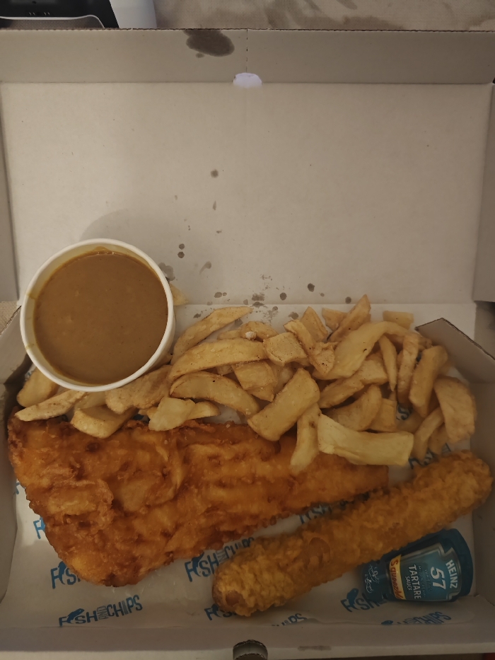 A cardboard box contains a takeaway meal of fish and chips. A large battered fish fillet sits alongside a fried sausage and a pot of gravy. A generous portion of chips covers part of the fish. A small bottle of Heinz Tartare Sauce is also included. The box interior is slightly greasy, and "FISH & CHIPS" is printed on the bottom left corner.