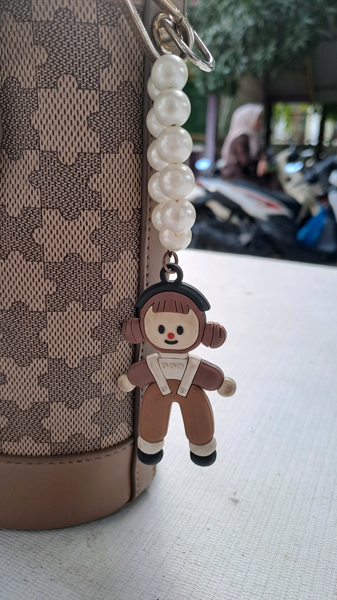 A close-up shot focuses on a purse with a patterned texture and a charm hanging from it. The charm is a small, cartoonish figure of a person with a brown hat, brown overalls, and white accents. A string of white pearls dangles above the charm. The purse is resting on a white, textured surface, possibly a table. In the blurred background, several motorcycles are visible, along with a person wearing a headscarf. The environment appears to be outdoors in Julicotmeurak, Indonesia.
