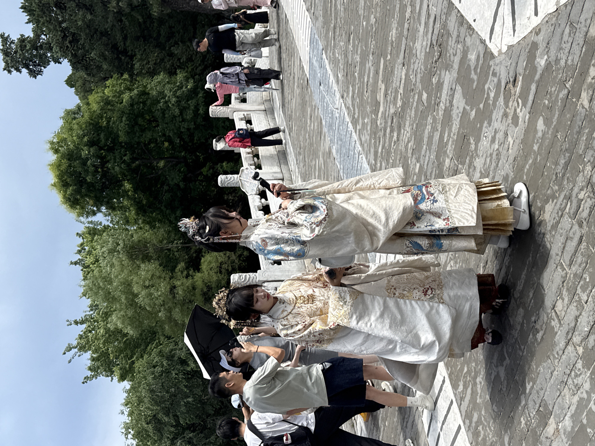 This image depicts an outdoor scene on a paved pathway or bridge within a park-like environment, located in Haidian District, China.
In the foreground, two young women are prominent, dressed in elaborate traditional Chinese attire. The woman on the left wears a cream-colored outer robe extensively embroidered with gold patterns of birds and foliage, over dark red undergarments. She has a complex gold headpiece and holds a piece of white fabric in her left hand while looking downwards. The woman on the right wears a similar cream-colored robe, featuring blue and red embroidered phoenix or dragon motifs on the sleeves and lower skirt, over a pleated gold undergarment. She has an intricate silver and gold headpiece with dangling ornaments and holds a small black object in her left hand, also looking downwards. Both women wear traditional-style footwear.
Behind them and to the left, several individuals in modern clothing are visible. A man in a light green t-shirt and dark shorts stands, holding a black umbrella over a woman in a grey dress, white cap, and sunglasses. Another man in a white t-shirt and black pants carries a backpack.
Further back, on a white stone bridge with ornate carved railings, additional people are present. One individual in a pink top leans against the railing, possibly holding a selfie stick. Another person in a grey hooded jacket stands nearby. A man in a black t-shirt and light grey pants walks towards the right, carrying a bag. A woman in a red jacket and a beige hat walks away from the camera on the right side of the bridge. A young child in a dark dress is partially visible behind the man in the black t-shirt.
The pathway is constructed of large, rectangular grey paving stones, some appearing wet or discolored. Dense green trees form the background, under an overcast or partly cloudy sky. The scene is brightly lit by natural daylight.