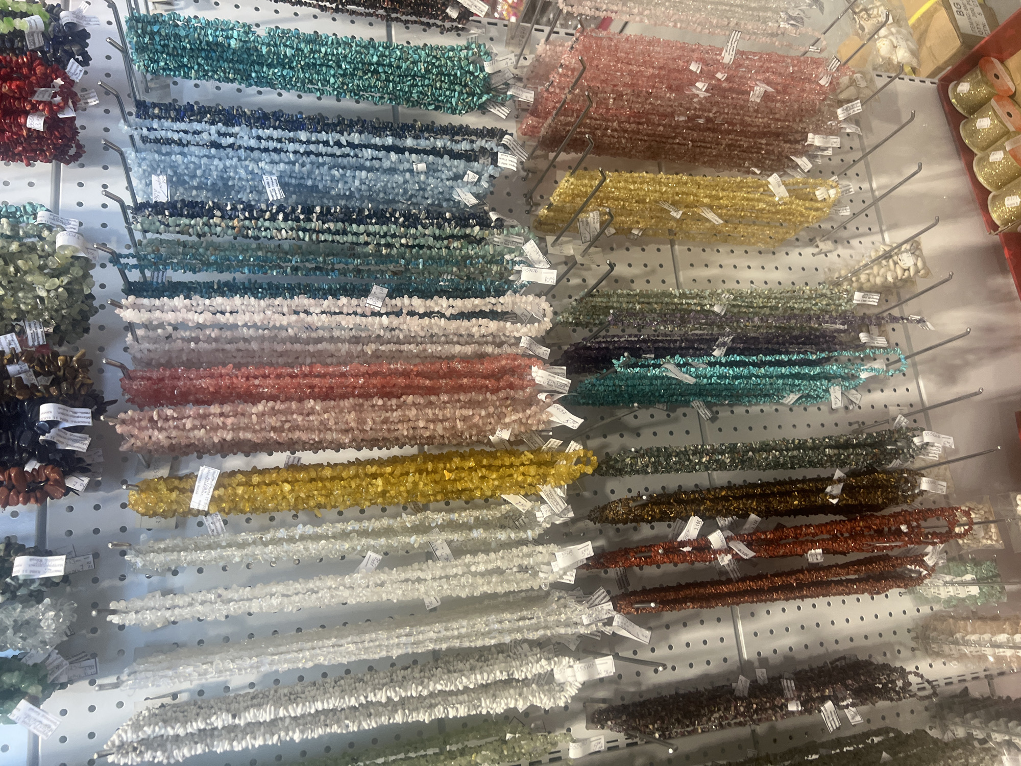 The image depicts a display of numerous strands of multicolored beads hanging on a white, perforated wall. The beads appear to be made of small, irregularly shaped gemstones or crystals in various colors including red, teal, blue, pink, yellow, green, brown, and white. Each strand is hung with a thin metal rod, and many have small white tags attached. In the upper right corner, there are packaged items with a red container in the background. Based on information from the tags the items are in Burgas, Bulgaria.