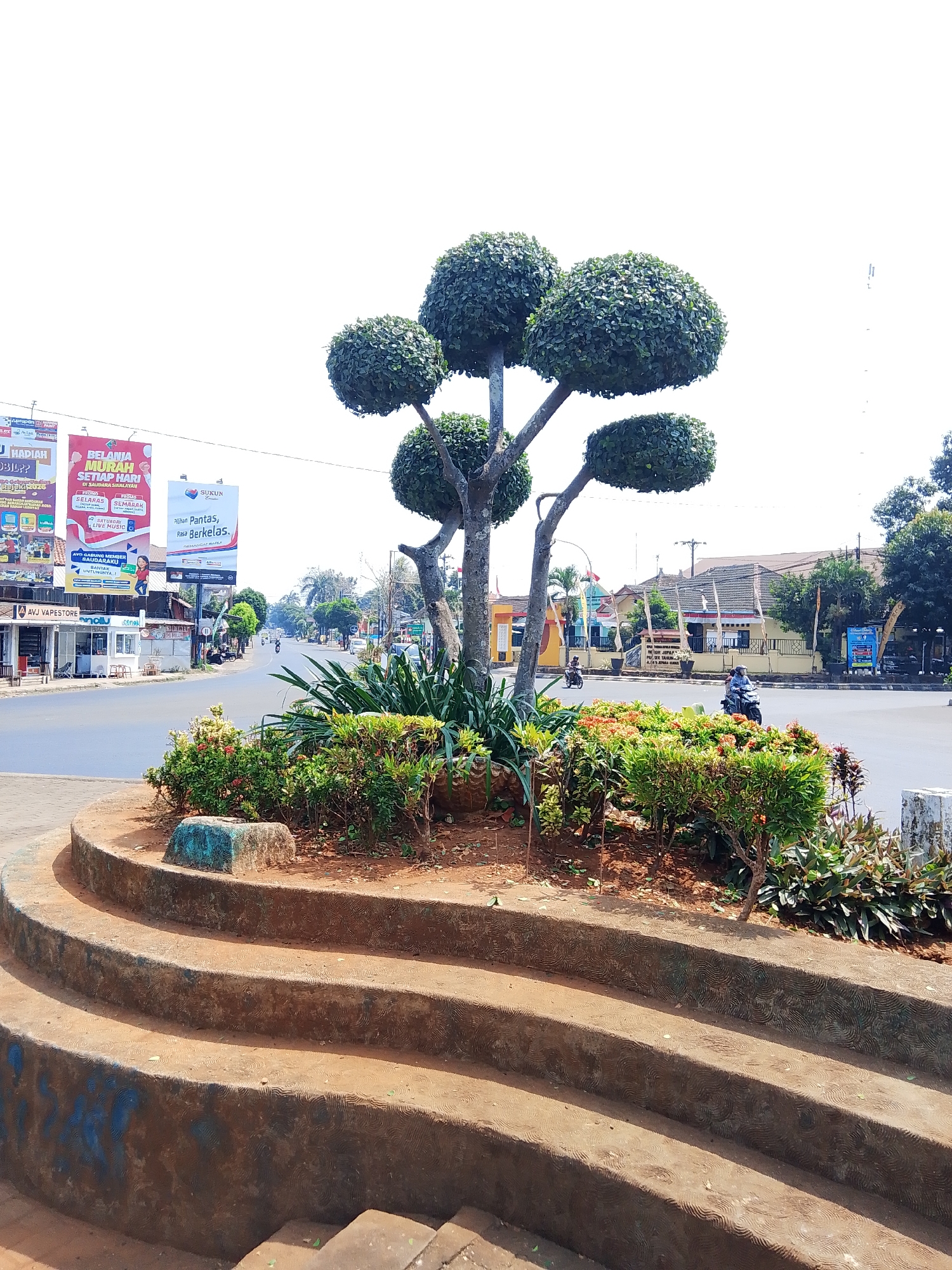 The image shows a street scene in Tahunan, Indonesia, with a prominent topiary tree in the foreground. The tree is sculpted into a spherical shape on a branching trunk. Behind the tree, a street curves to the left, lined with shops and buildings. Several billboards are visible on the left side of the street, advertising various products and services, including "BELANJA MURAH SETIAP HARI" and "SUKUN." A few motorcycles are seen on the street, with two riders. The environment is sunny with a bright sky. The foreground features a tiered circular planter made of concrete with a reddish-brown hue, decorated with blue paint splatters and a textured pattern. A decorative rock with a blue and green hue sits on the planter. Various low-lying green plants and some red-flowered bushes are planted within the planter. The background includes residential buildings with tiled roofs and dense foliage. The scene depicts a typical urban street with commercial establishments and residential areas, enhanced by a decorative landscape feature.