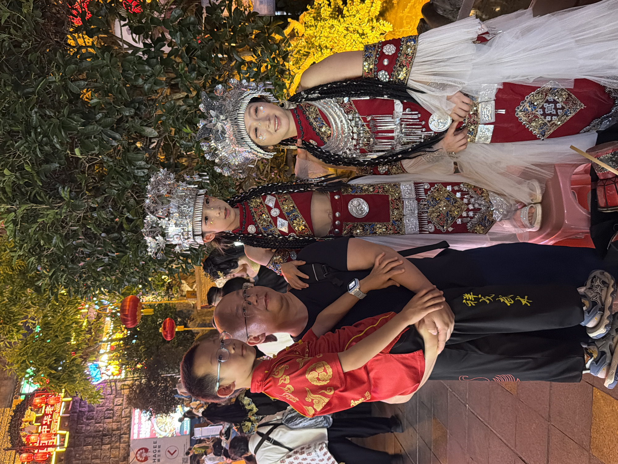 In Yuzhong District, China, a family of four is posing for a photograph at night, illuminated by festive red and yellow lights. The father, wearing a black t-shirt and black pants with yellow Chinese characters, is holding his young son, who is dressed in a traditional red and gold Chinese outfit. To the right of the father are two girls in elaborate traditional attire. Both girls are wearing intricate silver headdresses, red tops adorned with silver and gold embellishments, and long, white tulle skirts. The girl on the right has a silver necklace and earrings, and her face is decorated with glitter. The background features a stone wall, lush green trees, and hanging red lanterns, suggesting a cultural or celebratory event in an urban setting.