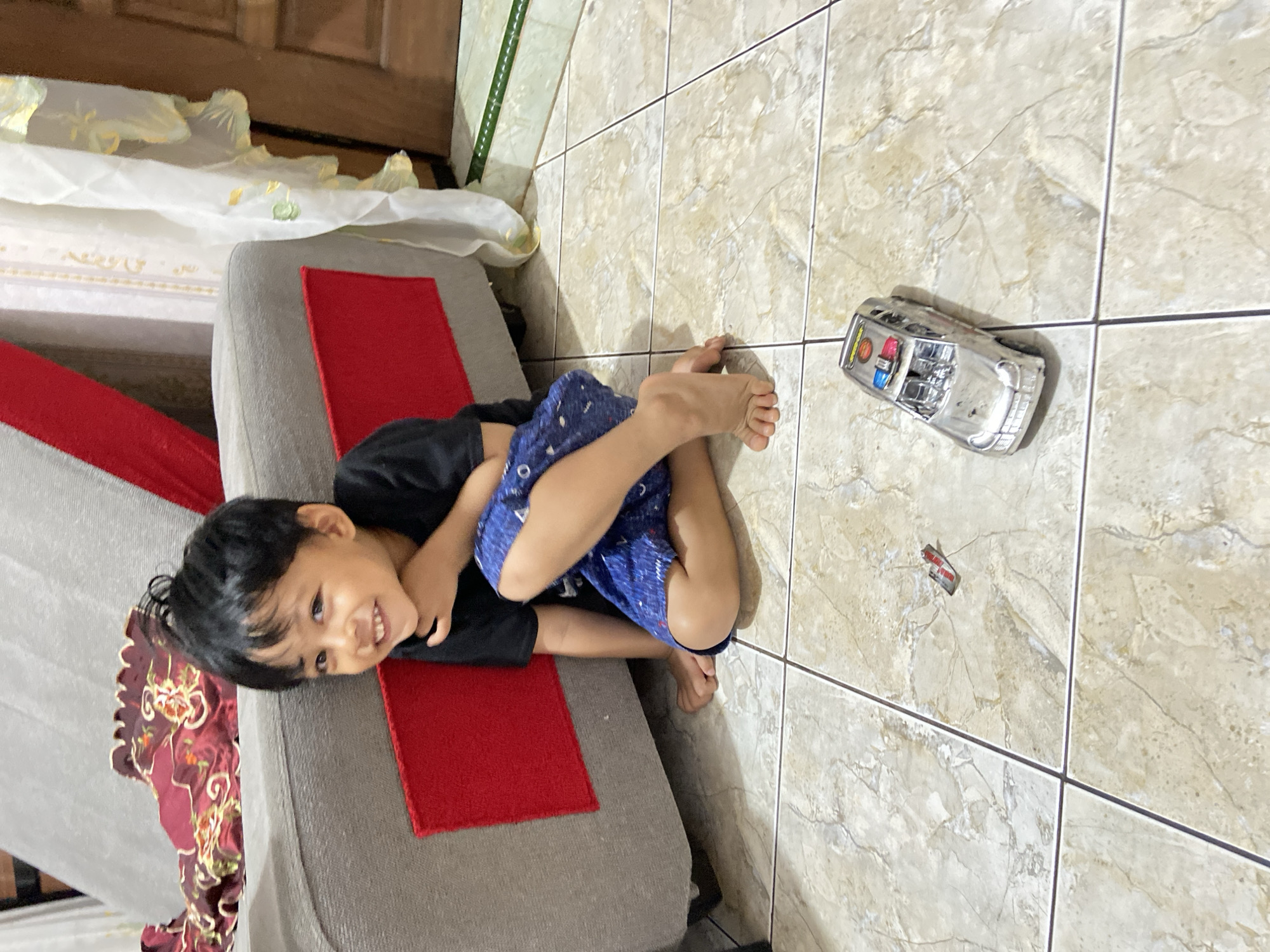 A young boy with black hair and a black t-shirt is sitting on a tiled floor in what appears to be a living room in Margorejo, Indonesia. He is wearing blue shorts with a white pattern. The boy is smiling and looking towards the camera, with his right hand resting on his chin. To his left, a grey sofa with red accents is visible. In front of him on the floor are two small toy vehicles, a silver police car with blue and red lights, and a smaller, indistinct red and silver object. A sheer, patterned curtain hangs on the right side of the frame, and a wooden door is partially visible behind it. The floor is tiled with a marble-like pattern.