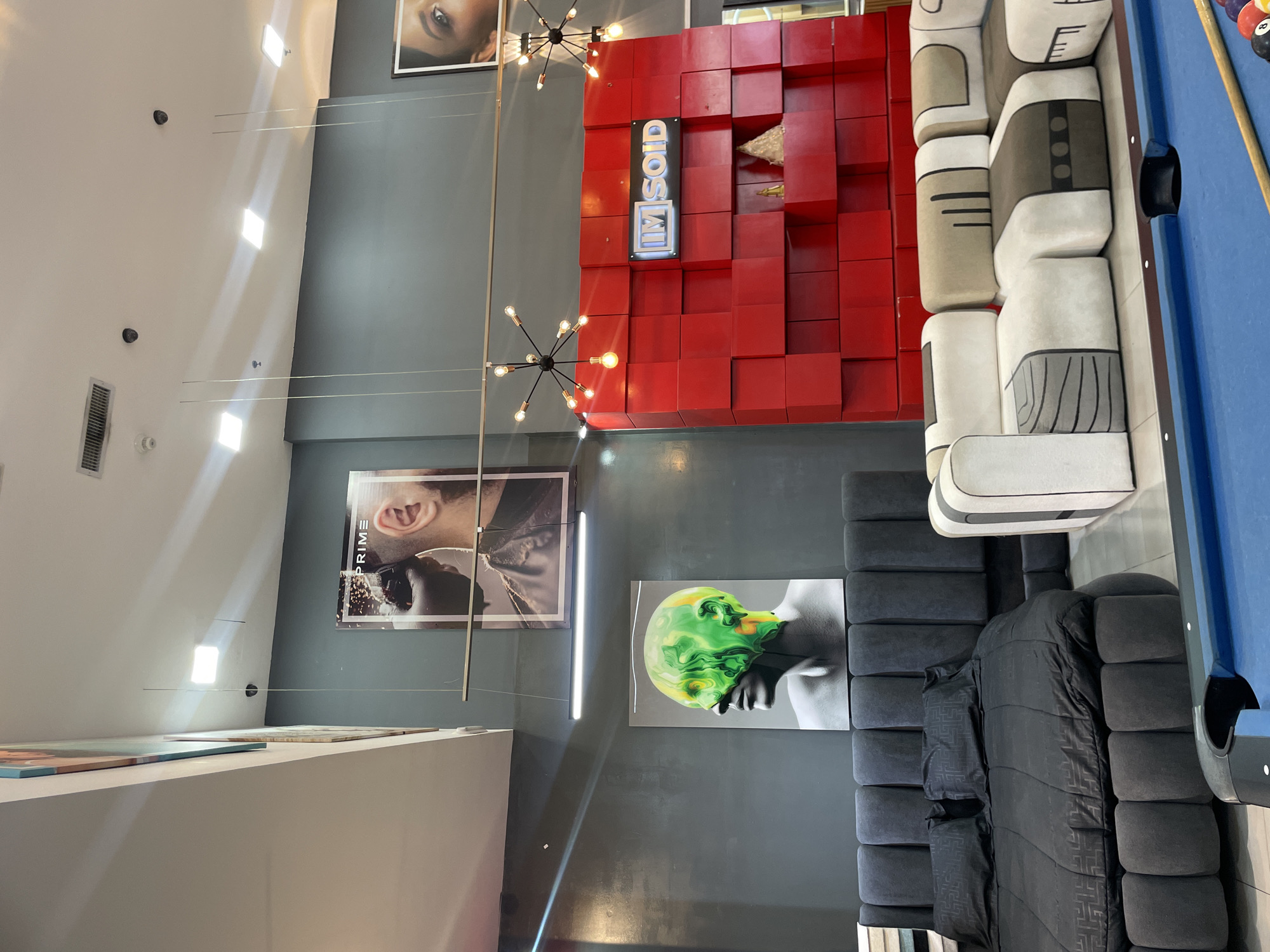The image depicts an interior scene, likely a furniture showroom, in Pampatar, Venezuela. A large, dark gray, tufted bed with black bedding is centrally located. To the right of the bed is a modern, modular sofa with white, beige, and brown upholstery. In front of the sofa and bed, a blue pool table is visible, with a cue stick and billiard balls.

The wall behind the furniture is painted a dark gray. Mounted on this wall are two prominent pieces of art. The upper piece is a framed poster featuring a close-up of a person's head and ear with the word "PRIME" visible. Below this, a larger artwork displays a stylized human head with vibrant green and yellow liquid-like patterns.

To the right of the artwork, a striking red wall composed of cubed panels dominates the background. A sign that reads "IM SOID" is mounted on this red wall. The ceiling is white, with several square fluorescent lights and modern starburst-shaped light fixtures hanging at various heights. The environment appears to be indoors, well-lit, and suggests a commercial space selling high-end furniture and possibly decor.