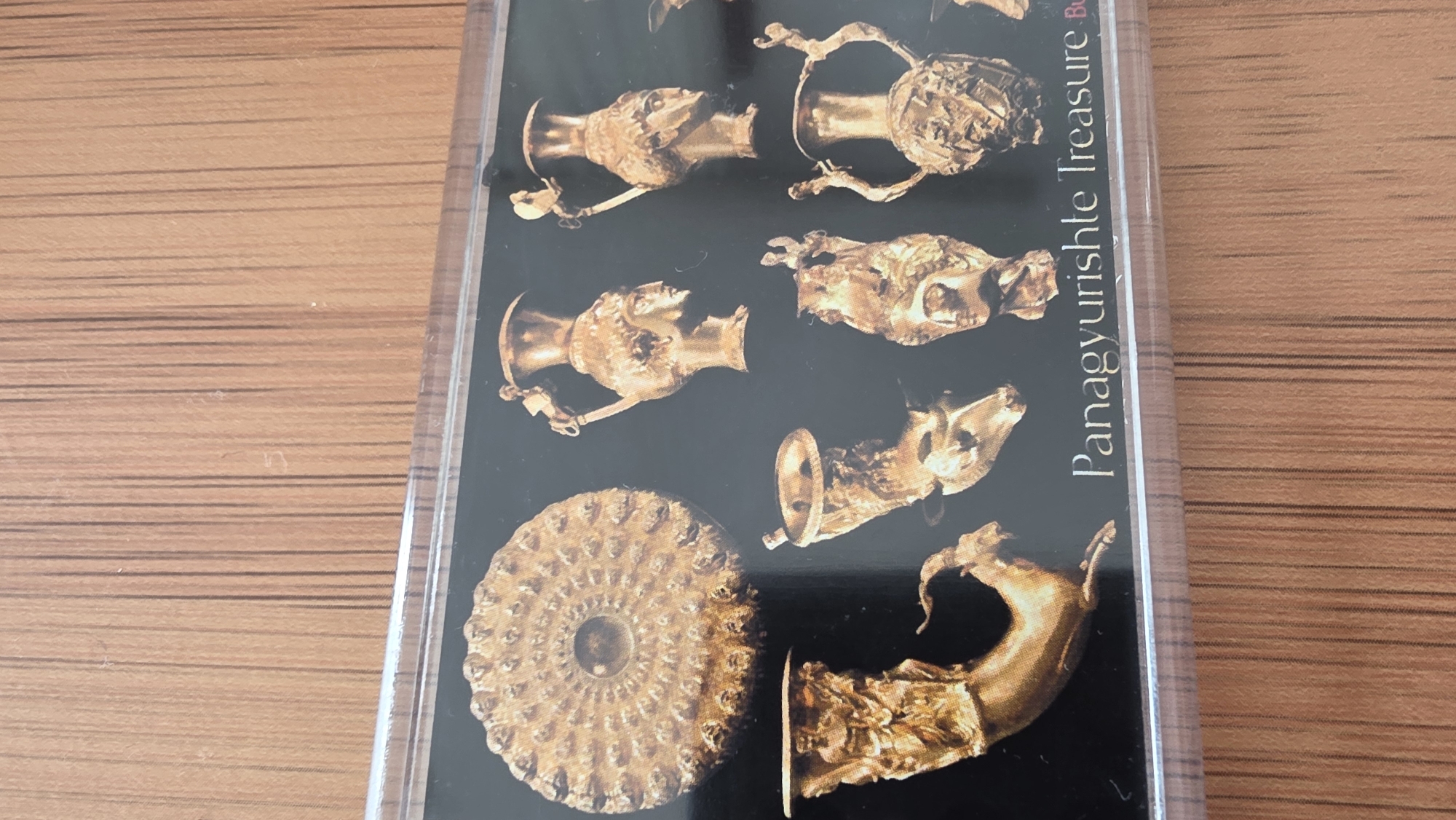 The image shows a pamphlet or card featuring a collection of golden artifacts, possibly ancient treasures, arranged against a black background. These artifacts include intricately designed vessels, a circular ornamented piece, and zoomorphic figures, some depicting animal heads or forms. The text "Panagyurishte Treasure" is visible on the right side of the image. The background is a wooden surface. There is no indication in this image that these objects are in Blagoevgrad, Bulgaria.