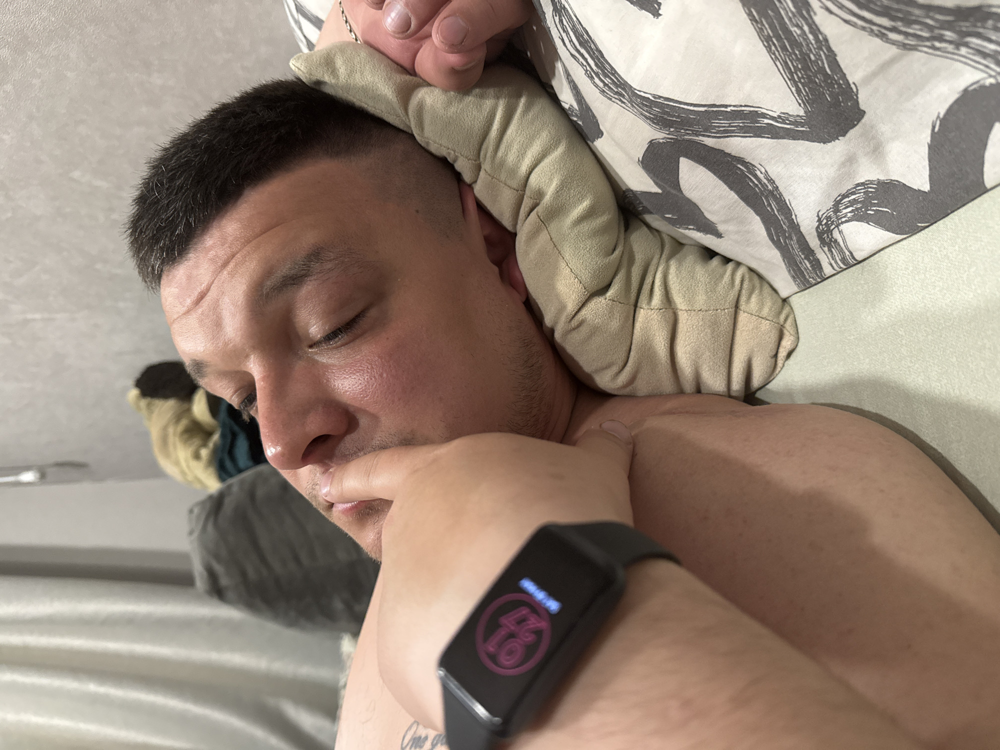 A man is lying down, presumably in a bed, with a pillow under his head. He has short, dark hair and his eyes are closed. He is wearing a black fitness tracker on his left wrist. The fitness tracker displays a pink graphic with numbers. A tattoo is partially visible on his arm. The background consists of a patterned pillow and bedding. The location in the city of Chaikovsky, Russia is not provided.