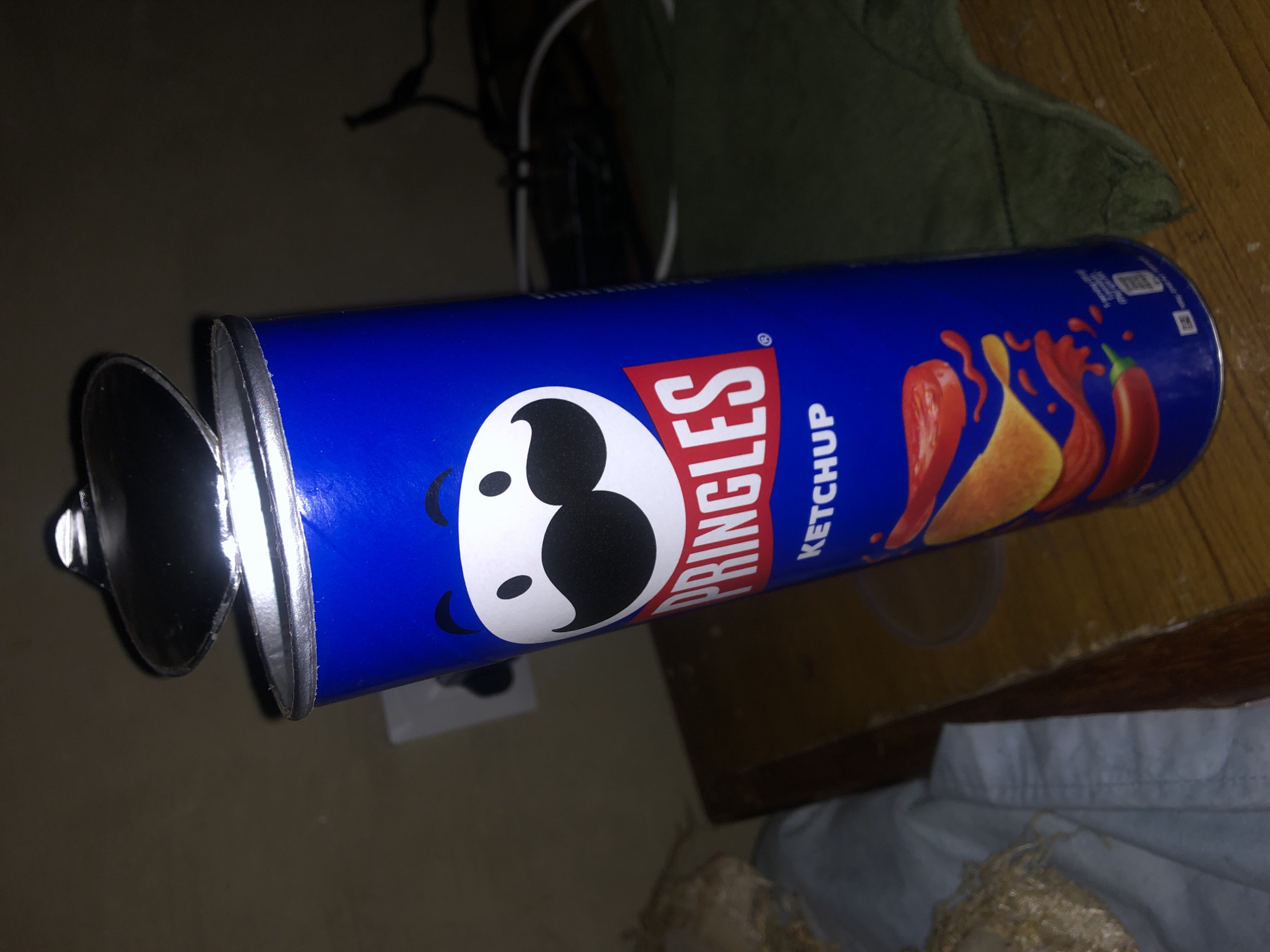 A Pringles Ketchup flavored potato chip can is shown in the foreground. The can is bright blue with the Pringles logo and a cartoon character of a face with a mustache. The lid of the can is partially open. The can is sitting on a wooden surface. To the left of the can, a white cloth object is partially visible. In the background, a wall with an electrical outlet and some dark wires are visible. The scene is dimly lit.