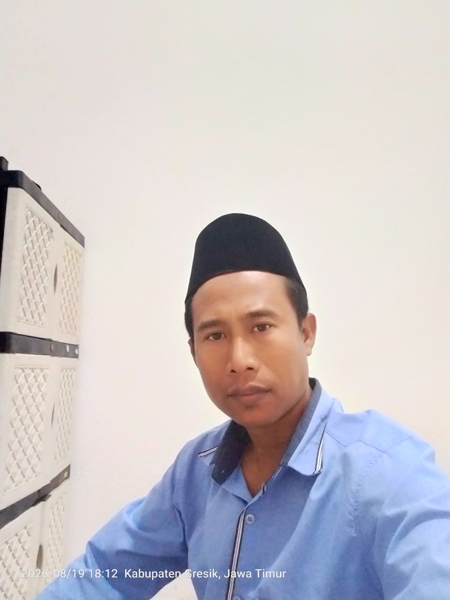 A man wearing a blue collared shirt and a black peci hat is looking at the camera. He is positioned to the right of a white cabinet with multiple drawers. The background is a plain white wall. A timestamp and location are visible in the bottom left corner of the image, reading "2026/08/19 18:12 Kabupaten Gresik, Jawa Timur".