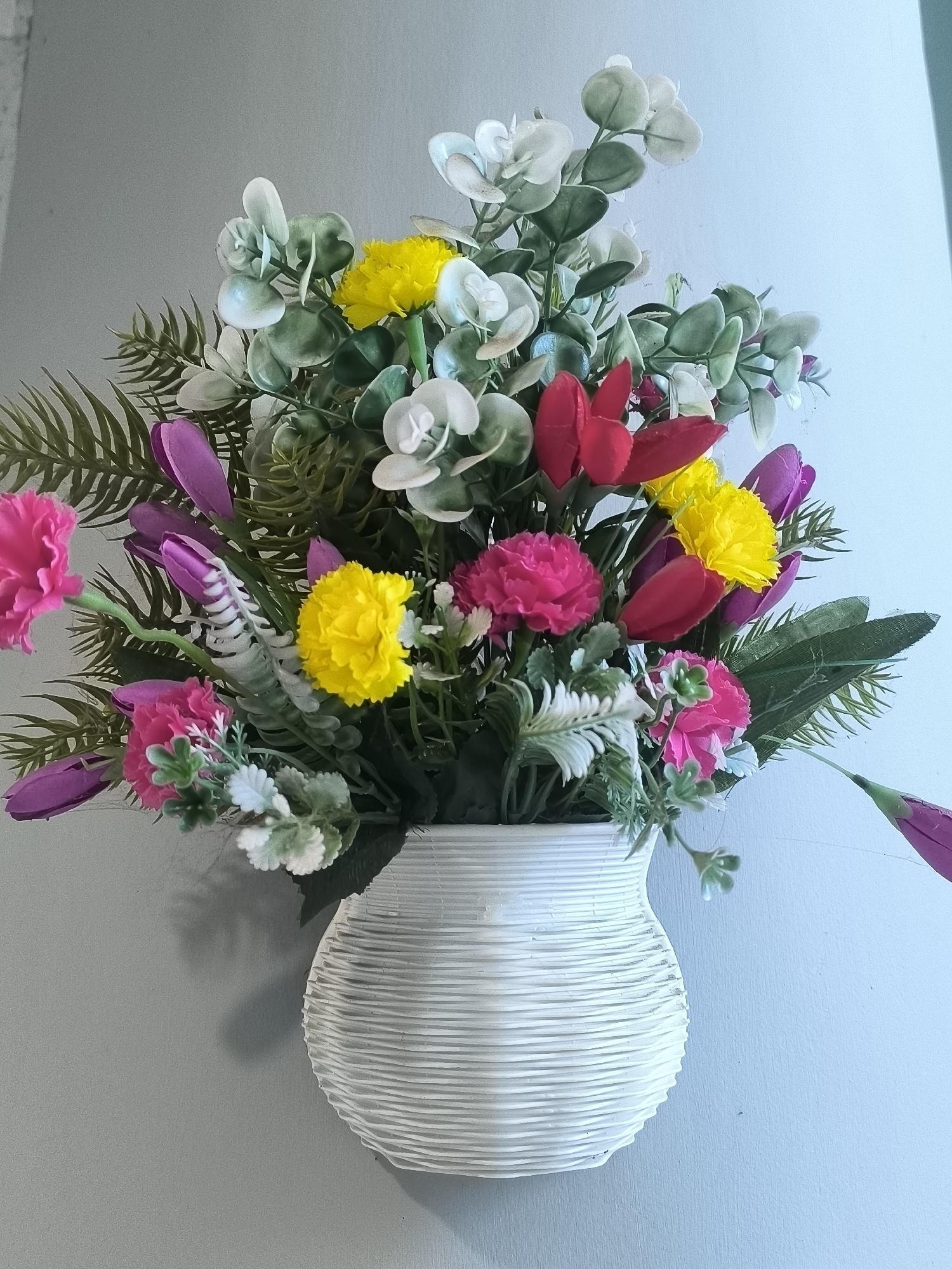 A vertical, eye-level shot captures a white, textured vase holding a vibrant arrangement of artificial flowers. The vase, adorned with horizontal ribbed lines, is positioned against a neutral gray wall. The bouquet itself is a dynamic composition of various artificial blooms and foliage in shades of yellow, pink, red, purple, and green. Prominent flowers include yellow carnations, pink carnations, red tulips, and purple tulip buds. Greenery, including what appears to be eucalyptus leaves and fern-like fronds, fills the spaces between the flowers, creating a lush and full display. The lighting casts a subtle shadow on the wall to the left of the vase, suggesting a light source from the right. The overall scene is clean and well-lit, with no visible people or indications of an outdoor environment. The background wall exhibits a slight gradient, shifting from a medium gray on the left to a lighter, bluish-gray on the right. This image is a still life composition, likely intended for decorative purposes, and its location within the city of Kalabbirang, Indonesia, is not visually indicated.