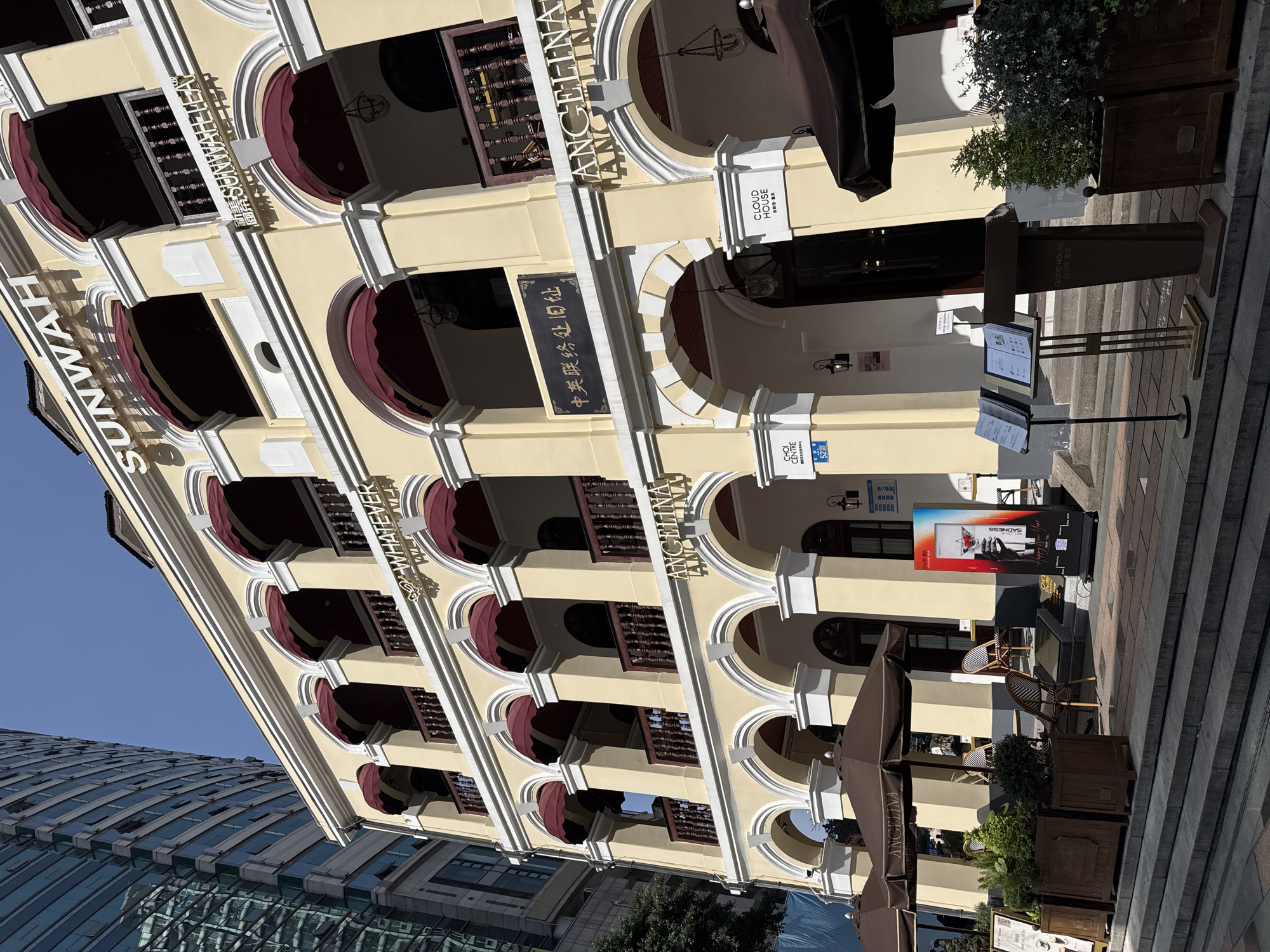 The image shows a building with multiple arches and balconies, featuring signage for "SUNWAH," "WHATEVER," and "ANGELINA." A sign with Chinese characters is also visible. The building is cream-colored with white trim and red awning-like structures over the arches. Street-side cafe tables and umbrellas bearing the "ANGELINA" name are present, along with signage for "CLOUD HOUSE" and "CHOI CENTRE 52." The modern glass-fronted building can be seen in the background. It's located in Yuzhong District, China.