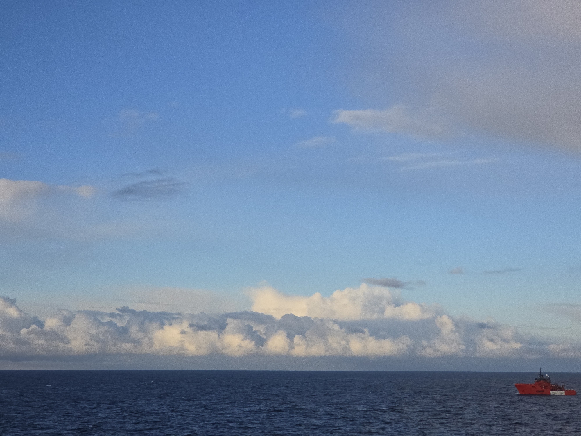 The image depicts an open ocean scene under a sky with varying cloud formations. The sky transitions from a clear blue at the top to a band of dense, cumulonimbus clouds along the horizon. A red vessel with white accents is visible on the right side of the frame, suggesting a possible maritime operation. The ocean appears dark blue, with gentle ripples on the surface. The location is Unknown, Unknown. There are no people visible in the image.