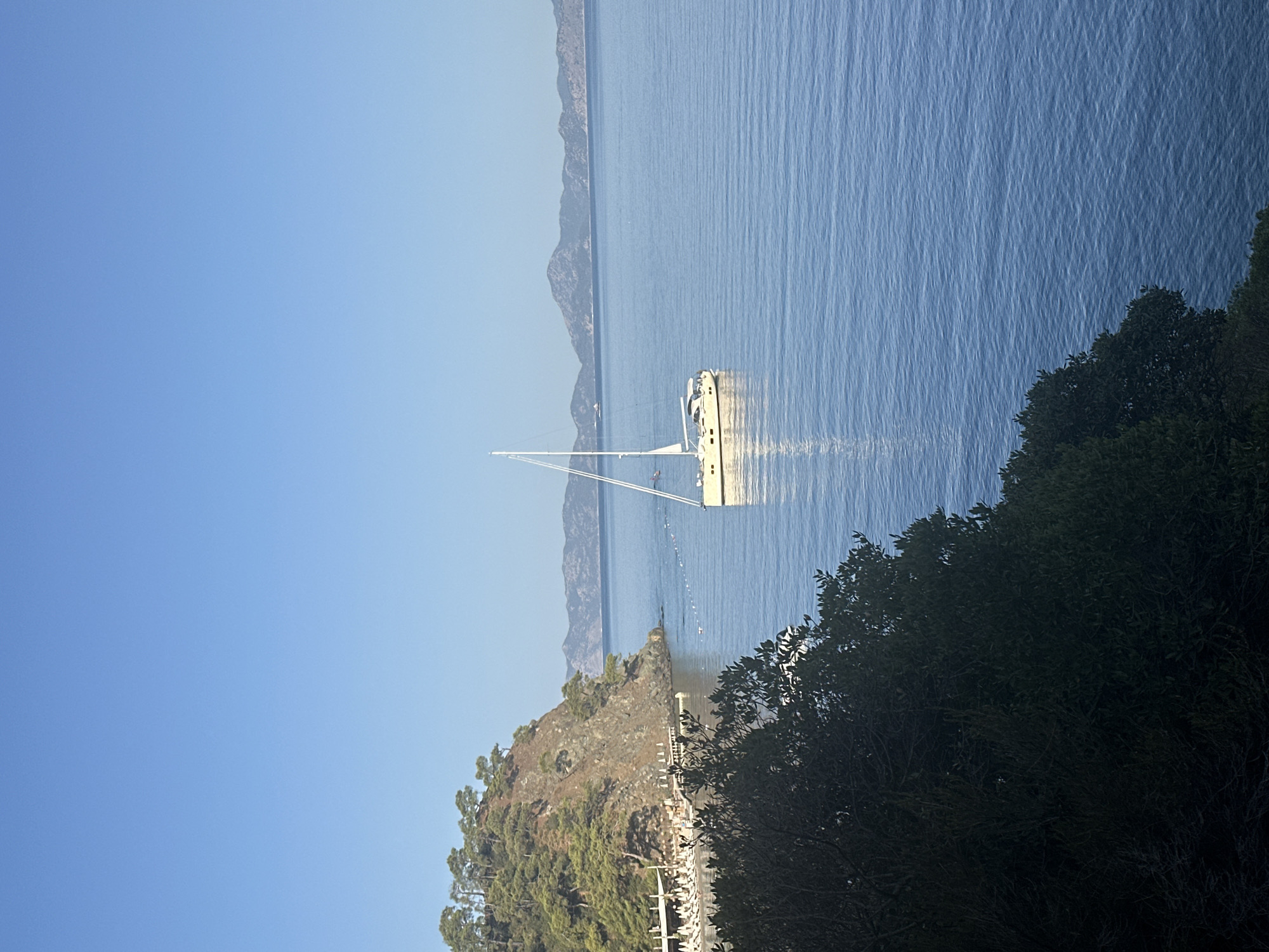 The image shows a picturesque coastal scene in Fethiye, Turkey, with a clear blue sky and calm, blue waters. In the foreground, dark green foliage frames the lower portion of the image. In the middle ground, a white sailboat with its mast tall and sails furled is anchored in the sea, gently reflecting on the water's surface. To the left, a steep, rocky hill covered in green pine trees rises from the coastline. Along the base of the hill, a beach area is visible with numerous white lounge chairs and umbrellas, suggesting a resort or public beach. A wooden pier extends into the water from the beach. In the background, a range of hazy, layered mountains are visible on the horizon, with a lighter shade of blue sky above them. The overall impression is one of a serene and beautiful natural landscape.