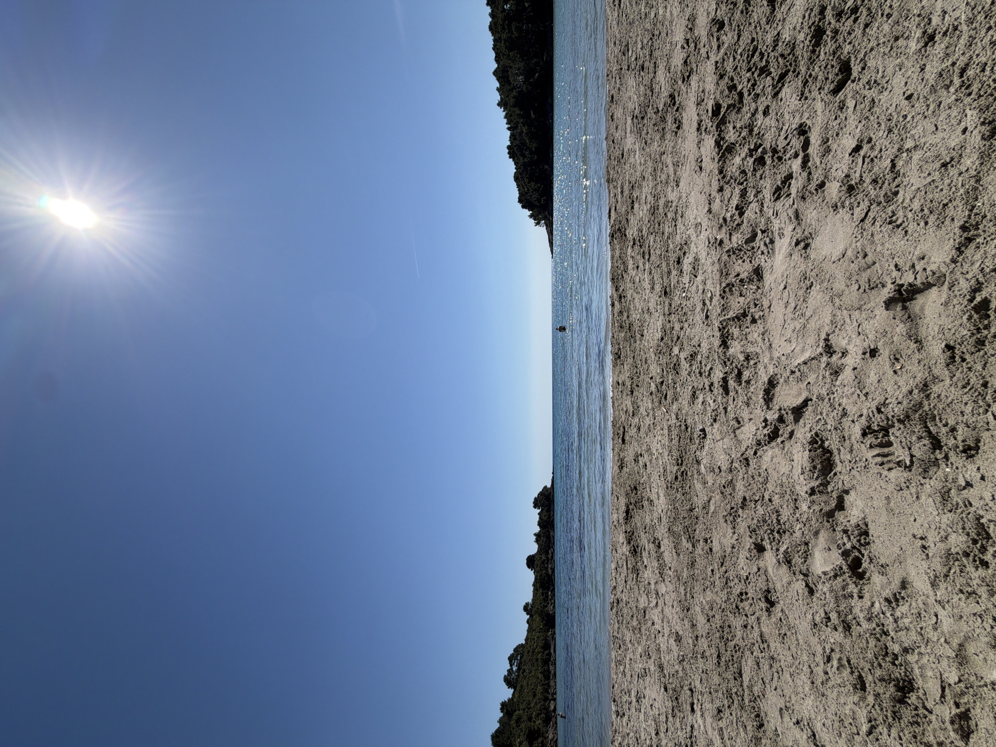 The image depicts a beach scene in Milovići, Montenegro, oriented with the horizon running vertically. A bright sun dominates the upper left corner of the sky. The scene includes a dark-colored sandy beach on the right side of the image and blue water and sky on the left. Vegetation appears to frame the upper and lower center of the water line. A dark, indistinct figure is visible in the water near the horizon. The sand shows textured patterns.