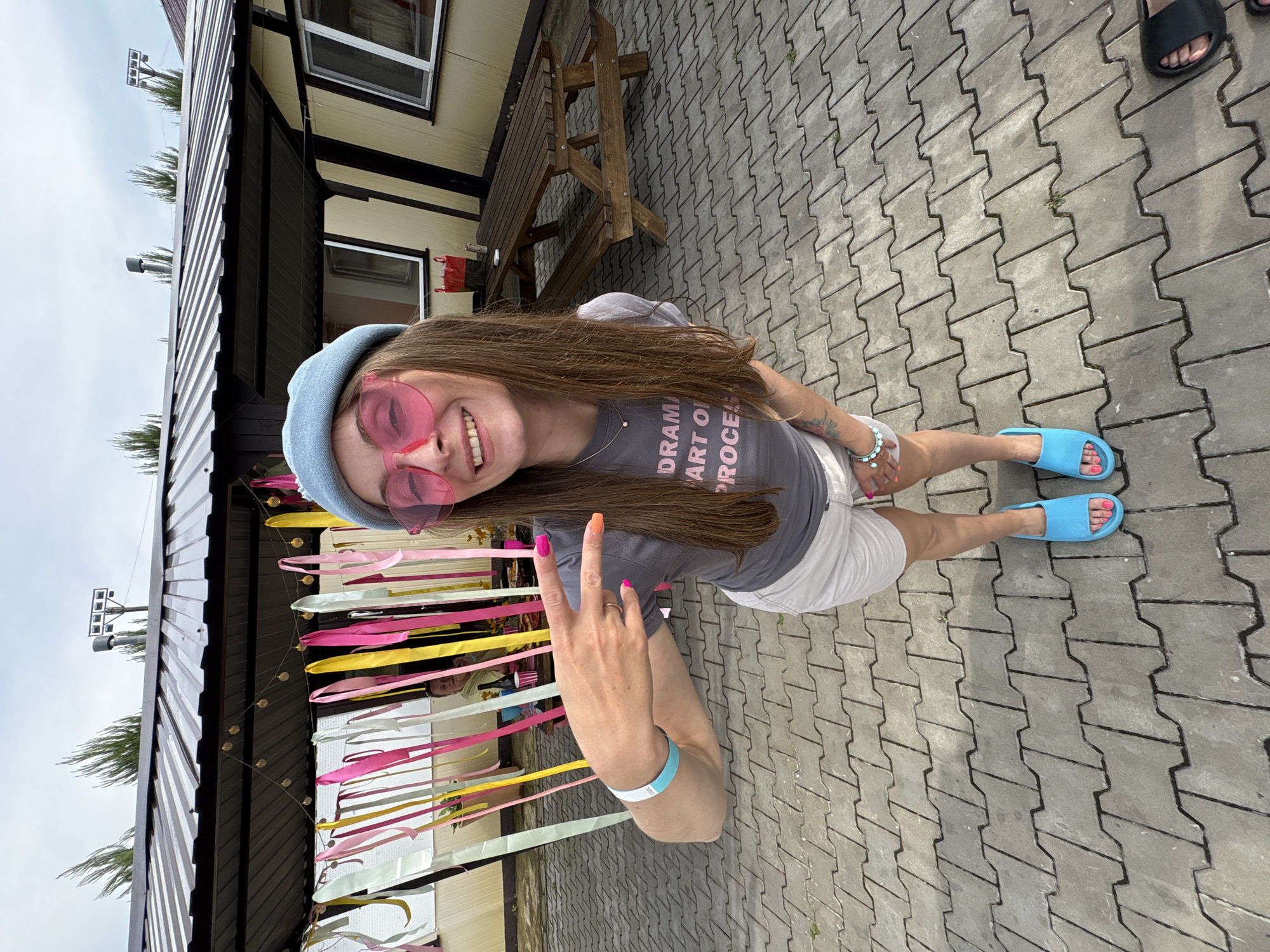 The image shows a young woman standing on a paved area in front of a building with a metal roof. She is wearing a light blue beret, large pink heart-shaped sunglasses, a grey t-shirt with pink lettering that reads "DRAMA IS PART OF PROCESS", white shorts, and blue slide sandals. She is making a "rock on" hand gesture with her right hand and is wearing a light blue wristband. Colorful streamers hang from the building behind her. A wooden picnic table and benches are visible to the right of the woman. The overall environment appears to be an outdoor event or recreational area, possibly in Birobidzhan, Russia, given the context.