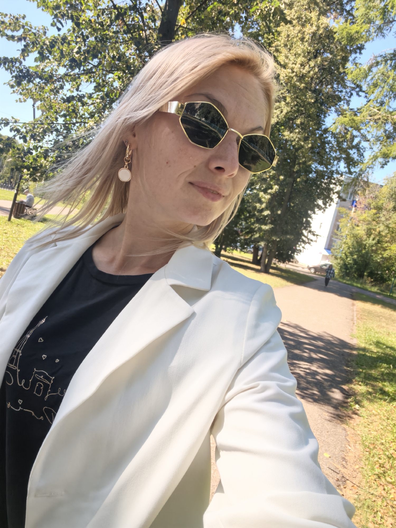 A woman with blonde hair and sunglasses is taking a selfie in an outdoor urban setting. She is wearing a white blazer over a black t-shirt with a graphic print. The background shows trees, a sidewalk, and a building. The image was taken on a sunny day.