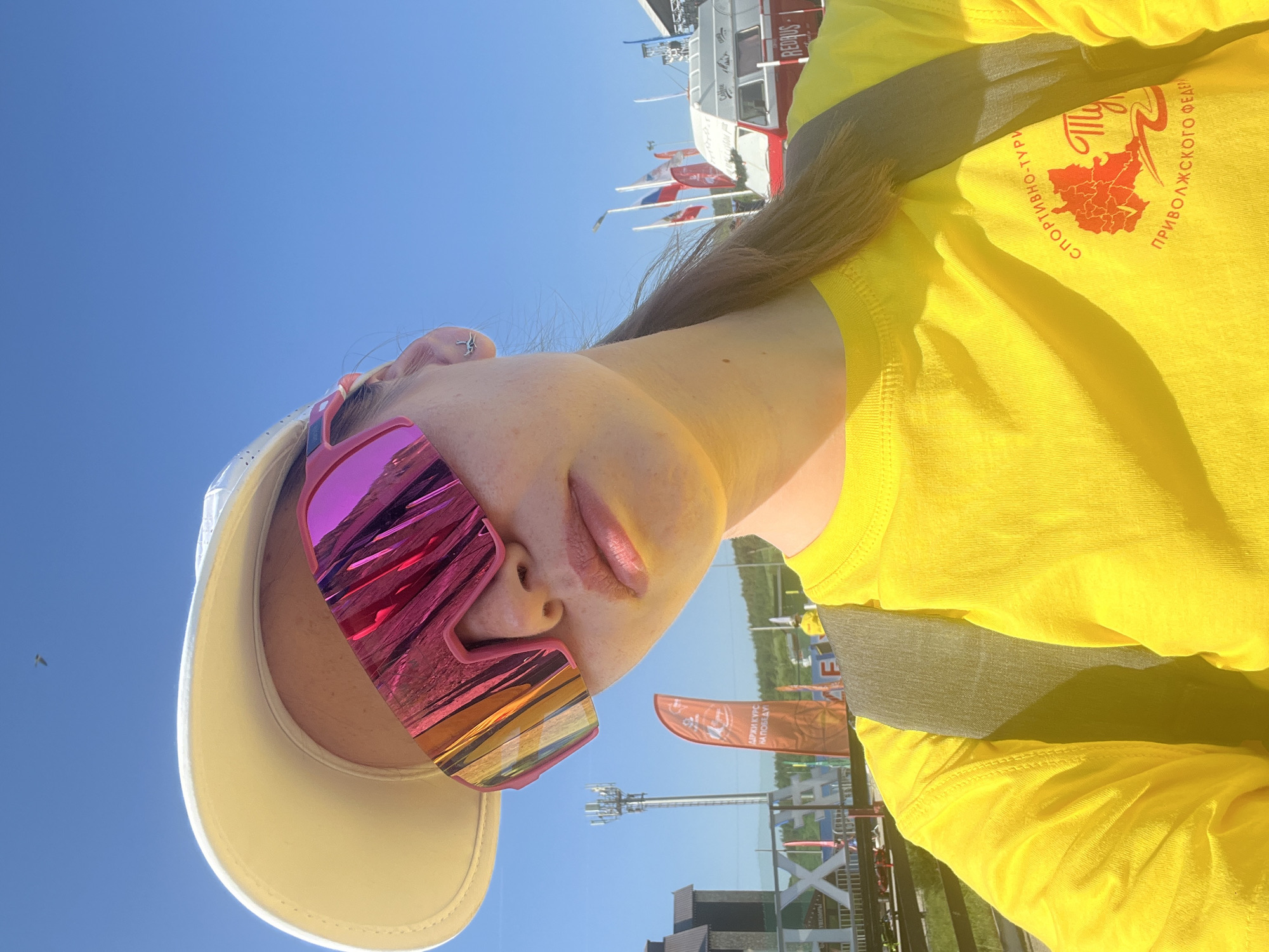 A woman in a yellow t-shirt and white baseball cap is wearing reflective sunglasses. The sky is blue, and there is a red and white bus in the background. The yellow t-shirt has a logo that says "Спортивно-Туристического Приволжского Феде". There are also flags and a tower in the background. This appears to be Khvalynsk, Russia.