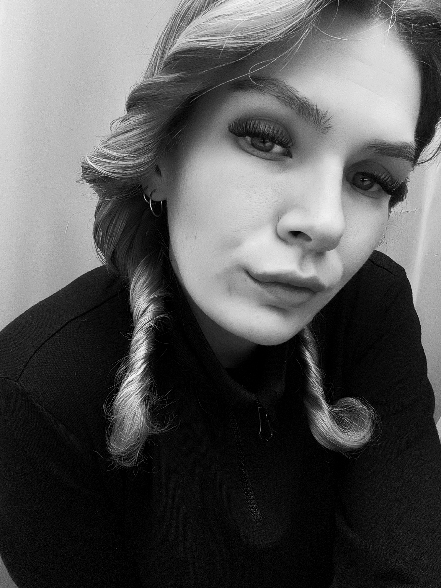 A black-and-white, close-up portrait shows a young woman with light-colored hair styled in twisted curls near her face. She has prominent eyebrows and long eyelashes, and wears small hoop earrings. She is wearing a dark, zip-up garment. The background is a plain wall.
The location Русский Вожой, Russia is irrelevant to the image content.