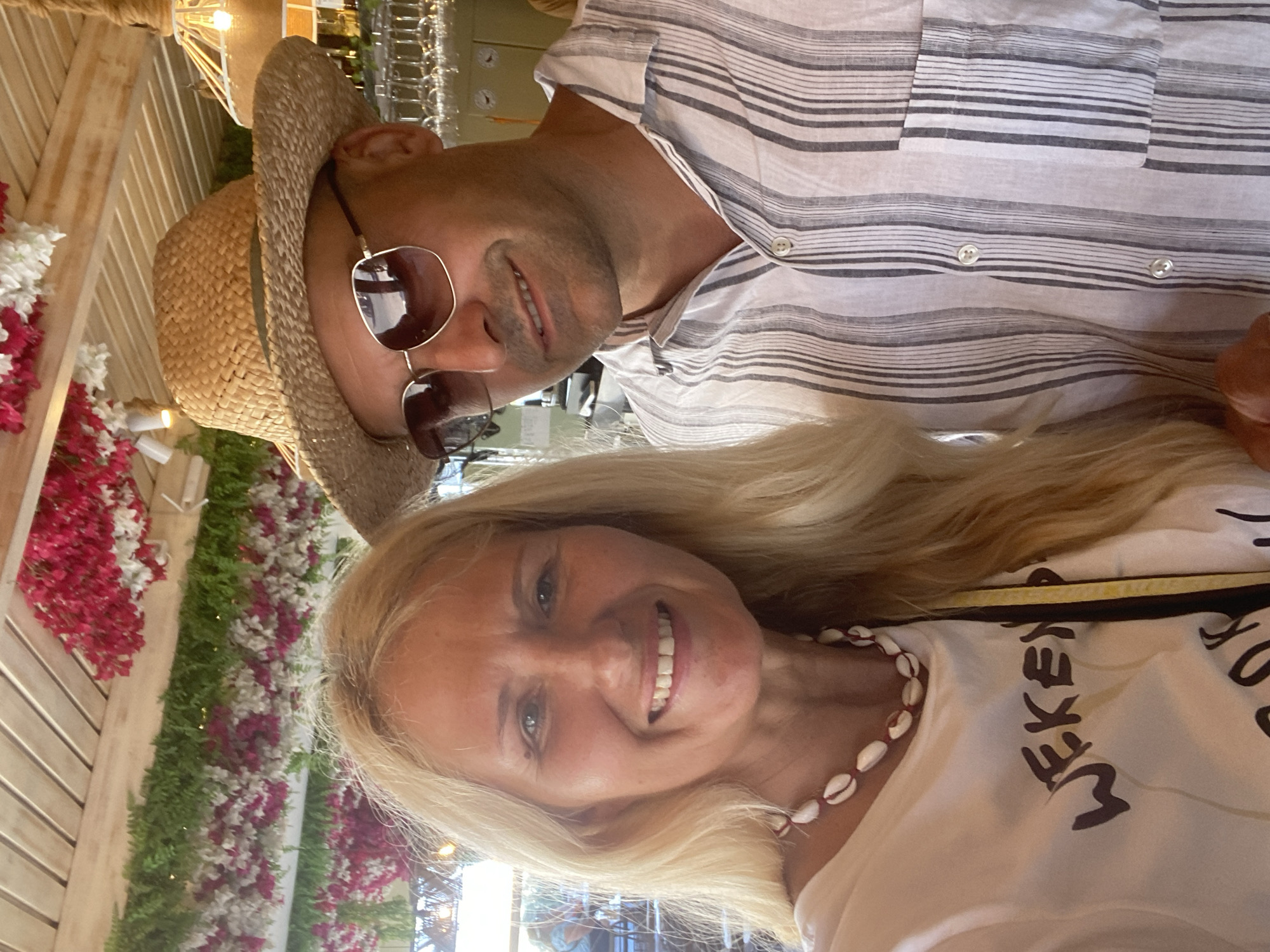 In an outdoor dining area with a wooden slatted ceiling adorned with hanging pink and white flowers, a blonde woman in a white t-shirt and a seashell necklace smiles at the camera. Beside her, a man wearing a straw hat and sunglasses, with a striped button-down shirt, also looks towards the camera. In the background, wine glasses are visible on shelves, suggesting a restaurant or bar setting, possibly in Ia, Municipal Unit, Greece.