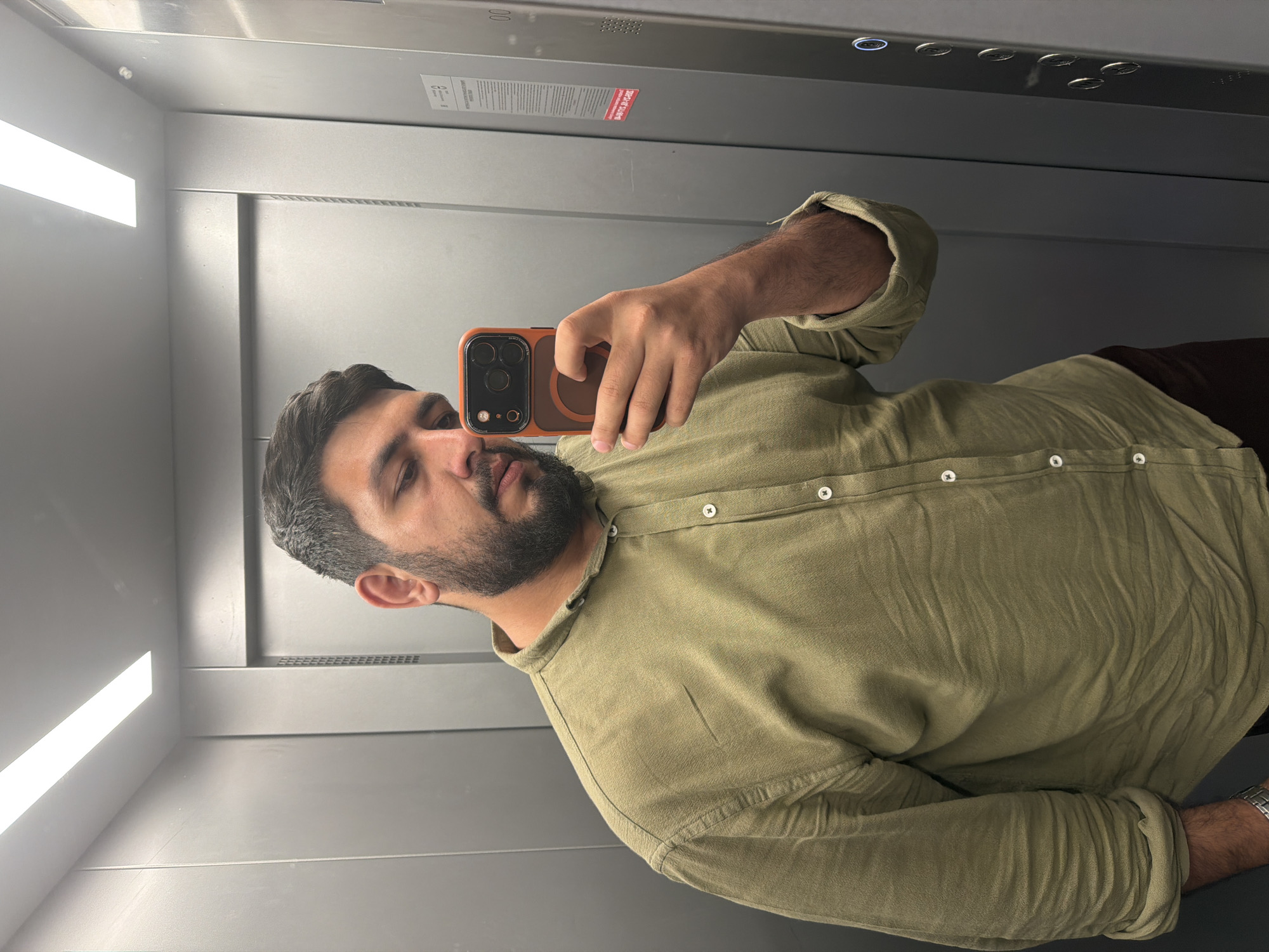The image displays a rotated selfie of a man taken inside an elevator. The man, appearing to be in his 20s or 30s with dark hair and a neatly trimmed beard, is wearing a light olive green long-sleeved collared shirt and dark trousers. He is holding an orange-brown smartphone with a distinct black camera module and a circular MagSafe ring on its back, capturing his reflection in the elevator's metallic wall. A silver-toned watch is visible on his left wrist.
The setting is a modern elevator interior with grey, reflective metallic walls that act as a mirror. Bright, rectangular strip lights are visible on the ceiling, casting illumination throughout the cabin. Above what would be the man's head if the image were upright, an elevator control panel is visible, featuring several circular buttons, some with blue backlights. Faint numbers "00" are discernible near the top left of this panel, likely indicating floor information.
Two informational stickers are present on the elevator wall:
1. A white rectangular sticker with black text and a pictogram (possibly depicting a person), partially legible, suggesting elevator usage rules (e.g., "Правила пользования лифтом" - Elevator usage rules).
2. A red rectangular warning sticker with white text, rotated and slightly blurred. Visible text includes "ОПАСНО" (DANGER) and potentially "при пожаре" (in case of fire), indicating emergency or safety instructions.
The image captures a casual, everyday activity of taking a mirror selfie. The indoor setting does not provide cues about the time of day or weather. This elevator is located in Krasnoyarsk, Russia.