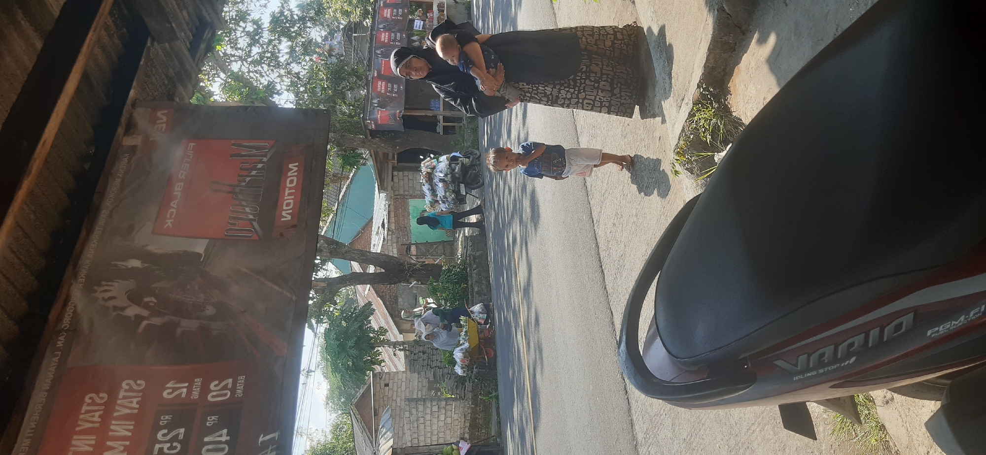 A street scene in Sepakek, Indonesia, captures a mother in a black hijab holding a baby. Beside her, a young boy in a blue t-shirt and white shorts stands on the sidewalk, looking at the camera. In the background, two women are visible at a market stall laden with produce, situated near a brick wall and trees. A motorcycle is parked behind them, also loaded with goods. The scene is framed by a banner advertising Marlboro cigarettes hanging overhead. The overall environment suggests a sunny day in a residential or market area, with typical street elements like asphalt roads and various structures.