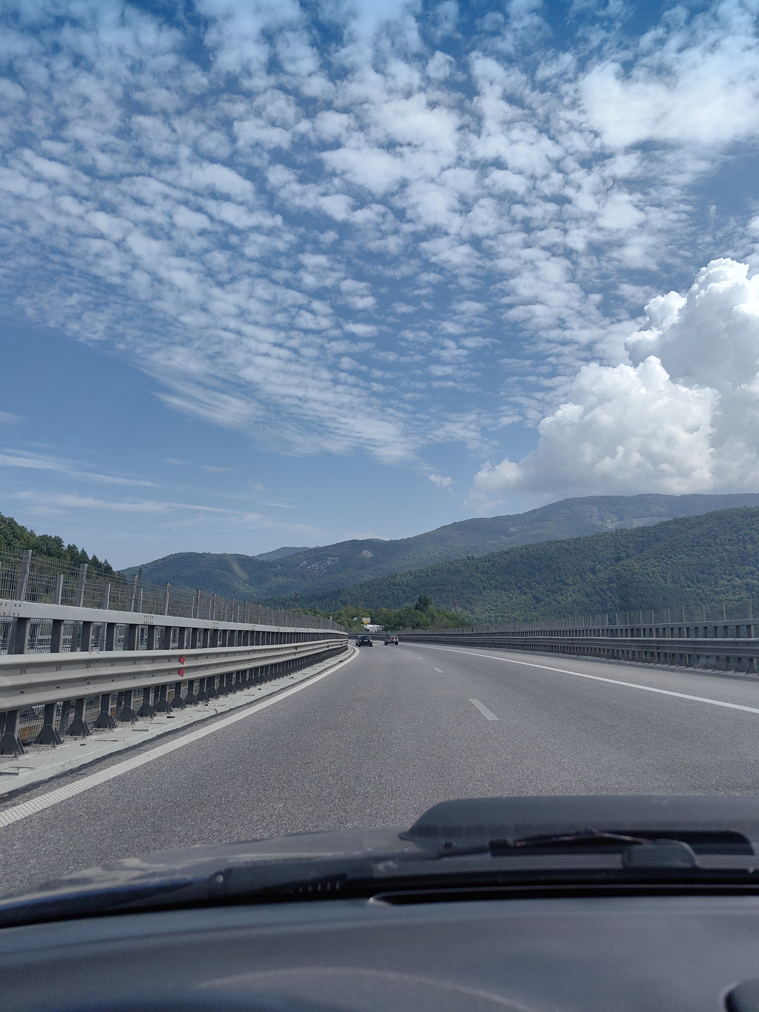 The image depicts a view from inside a vehicle traveling on a highway. The road is asphalt with a white lane marker visible. On the left side of the road, there is a metal guardrail with a fence behind it, and beyond that, a dense green forest covers the slopes of rolling hills. The sky is blue with scattered white clouds, some of which are large and puffy. In the distance, more tree-covered mountains are visible. Two vehicles are seen on the highway ahead, moving in the same direction as the camera's viewpoint. The dashboard of the vehicle, including the windshield wipers, is visible in the foreground. The scene appears to be in the region of Vrachesh, Bulgaria, suggesting a scenic route through a mountainous, forested area.