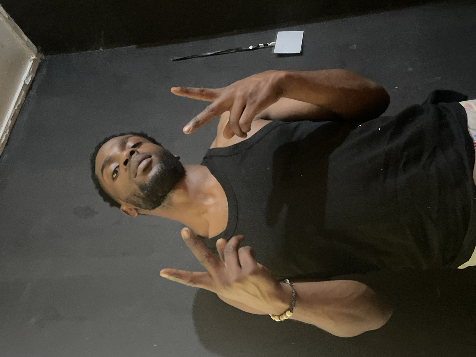 An adult male lies supine on a dark, possibly black, floor. He wears a black tank top and a bracelet on his right wrist. He holds up two fingers in the shape of a “V” with both hands. Above him, a white rectangular object is affixed to a black wall. It is not possible to determine the location of the image as Jos, Nigeria, with only the information provided.
