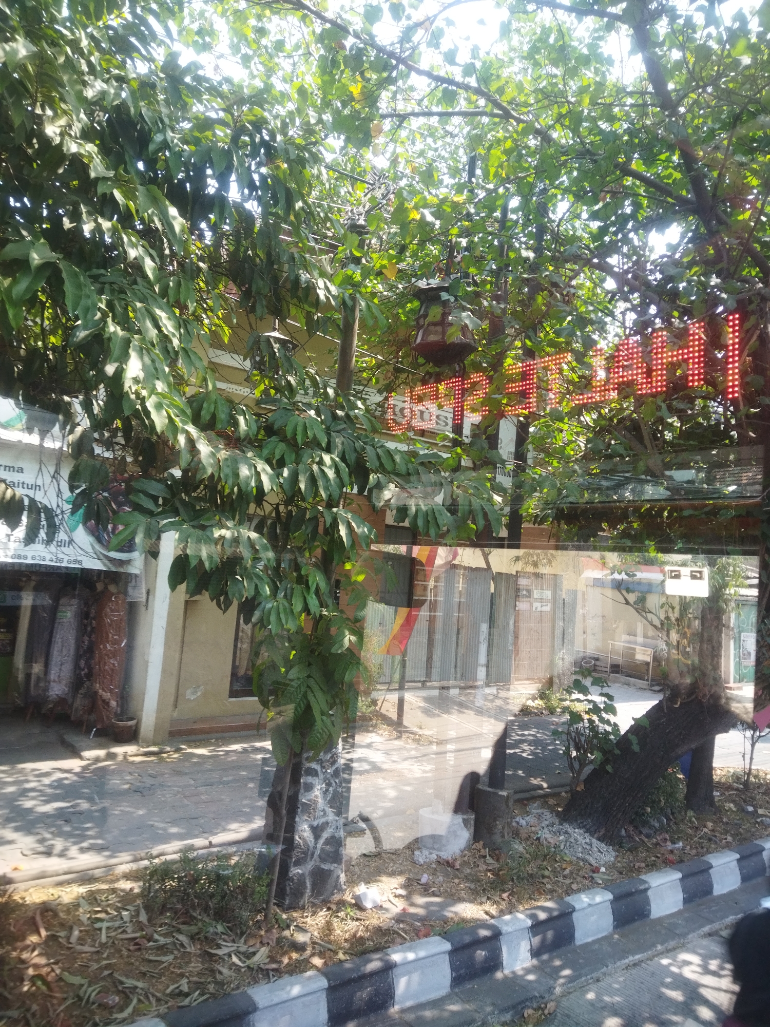 The image shows a street scene in Surakarta, Indonesia, on a sunny day. The foreground features a black and white striped curb and sidewalk. Beyond the curb, there is a patch of dry earth with scattered leaves and a small bush. A tree trunk with a stone-like texture is visible in the middle ground. In the background, there is a building with a storefront, some of which is obscured by trees. A red LED sign is visible above the storefront, reflecting in a glass pane. The leaves of the trees create a dappled shadow effect on the ground. The building appears to be a shop or business, with corrugated metal shutters on some of the entrances.