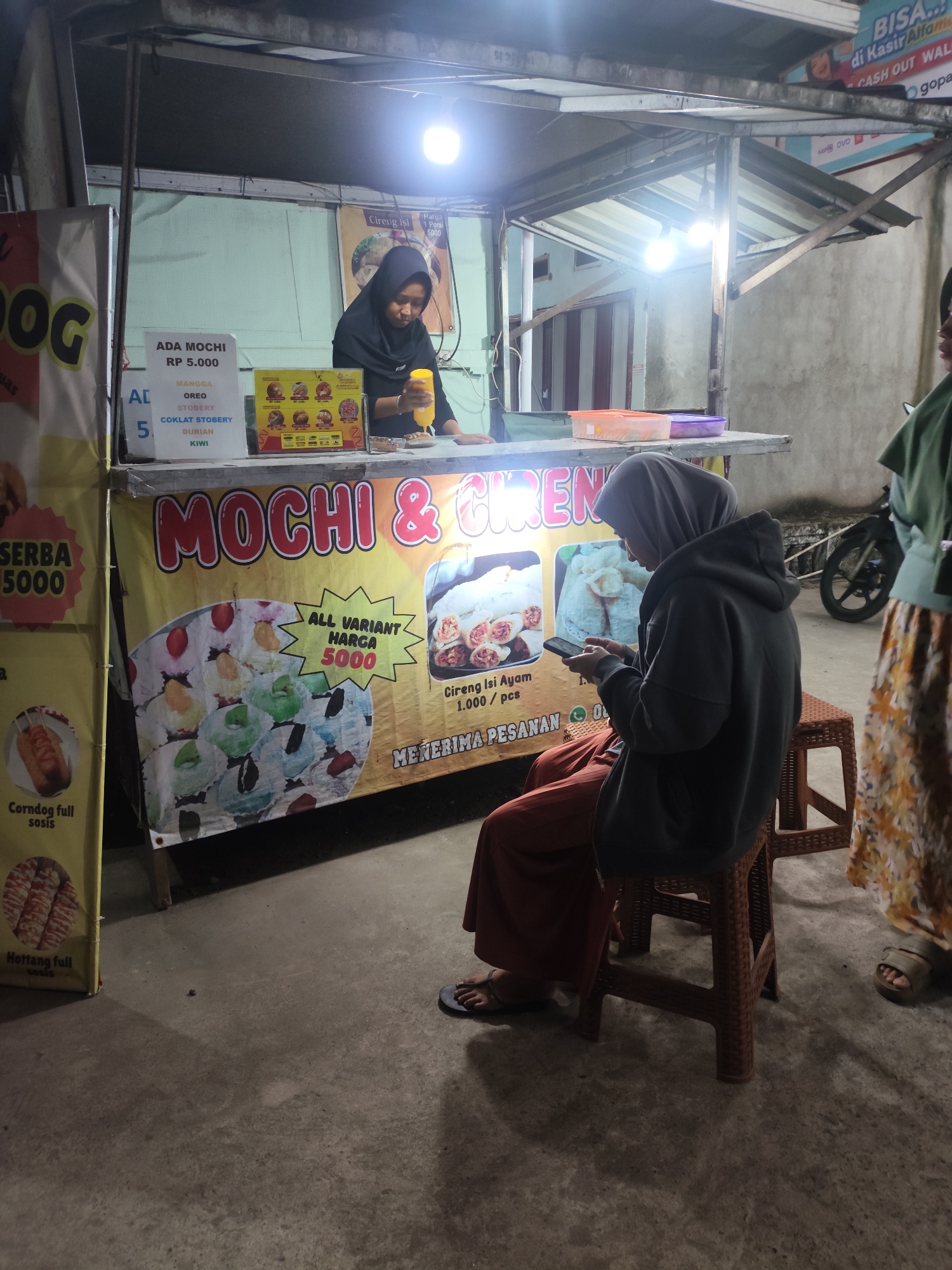 This is a medium shot, street-level, outdoor photograph taken at night in Palembang, Indonesia. The scene depicts a food stall selling mochi and cireng. In the background, a woman wearing a black hijab and black clothing is preparing food, holding a yellow squeeze bottle. To her right, a customer, also wearing a hijab and dark hooded jacket over an orange skirt, is seated on a brown plastic stool, looking at her mobile phone. She is wearing black flip-flops. Another person, wearing a green top and a patterned skirt, stands on the right side of the frame, partially visible. The food stall has a banner with text that includes "MOCHI & CIRENG" and images of the food items. A price list on the left indicates "ADA MOCHI RP 5.000" and lists various flavors like Oreo, Stobery, Coklat Stobery, Durian, and Kiwi. The stall also advertises "Corndog full sosis" and "Hettang full sosis". The ground is concrete and appears damp. A motorcycle is parked behind the stall. The lighting is artificial, with two bright bulbs illuminating the stall area. A sign with text including "BIISA..." and "di Kasir Alfam..." is visible above the stall.