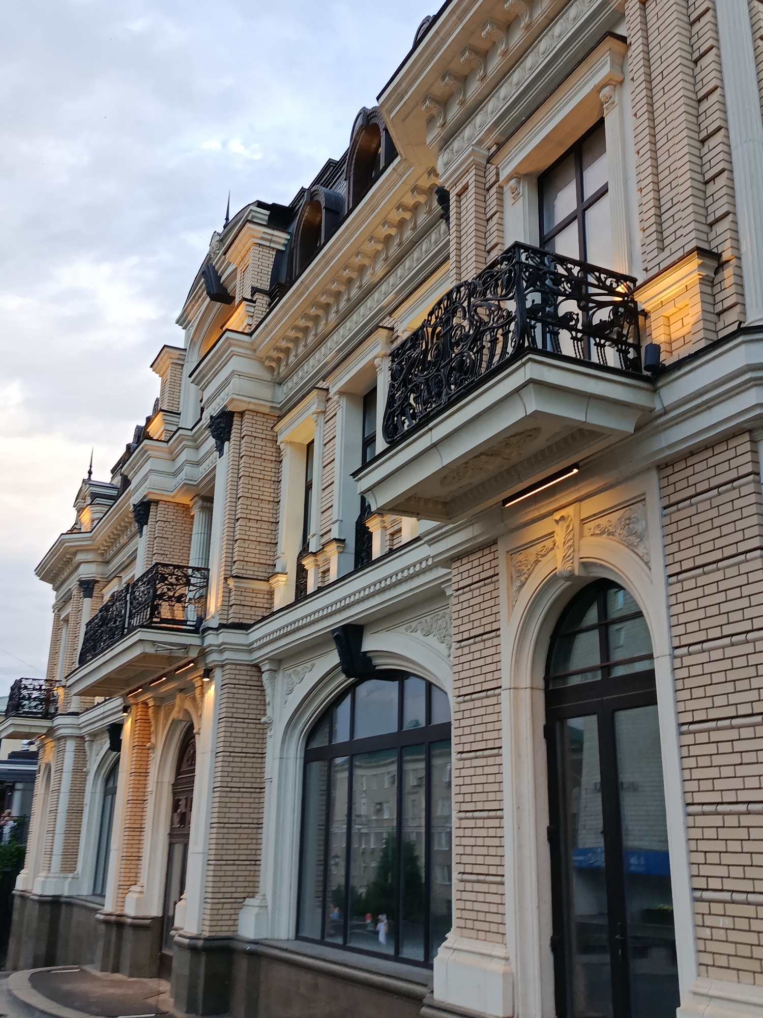 The image displays the exterior of a multi-story building, likely a hotel or residential complex, in Saratov, Russia. The architecture features light-colored brickwork, ornate white trim, arched windows, and dark balconies with intricate metalwork. The building has multiple levels, with several balconies visible on the upper floors. Artificial lighting illuminates some of the facade, highlighting the architectural details. The sky is overcast, suggesting it might be evening or a cloudy day. Reflections in the windows show other buildings, indicating an urban setting. No people are clearly visible in the foreground, though faint figures can be discerned in the reflections on the lower windows. The building appears well-maintained and is situated on a street.