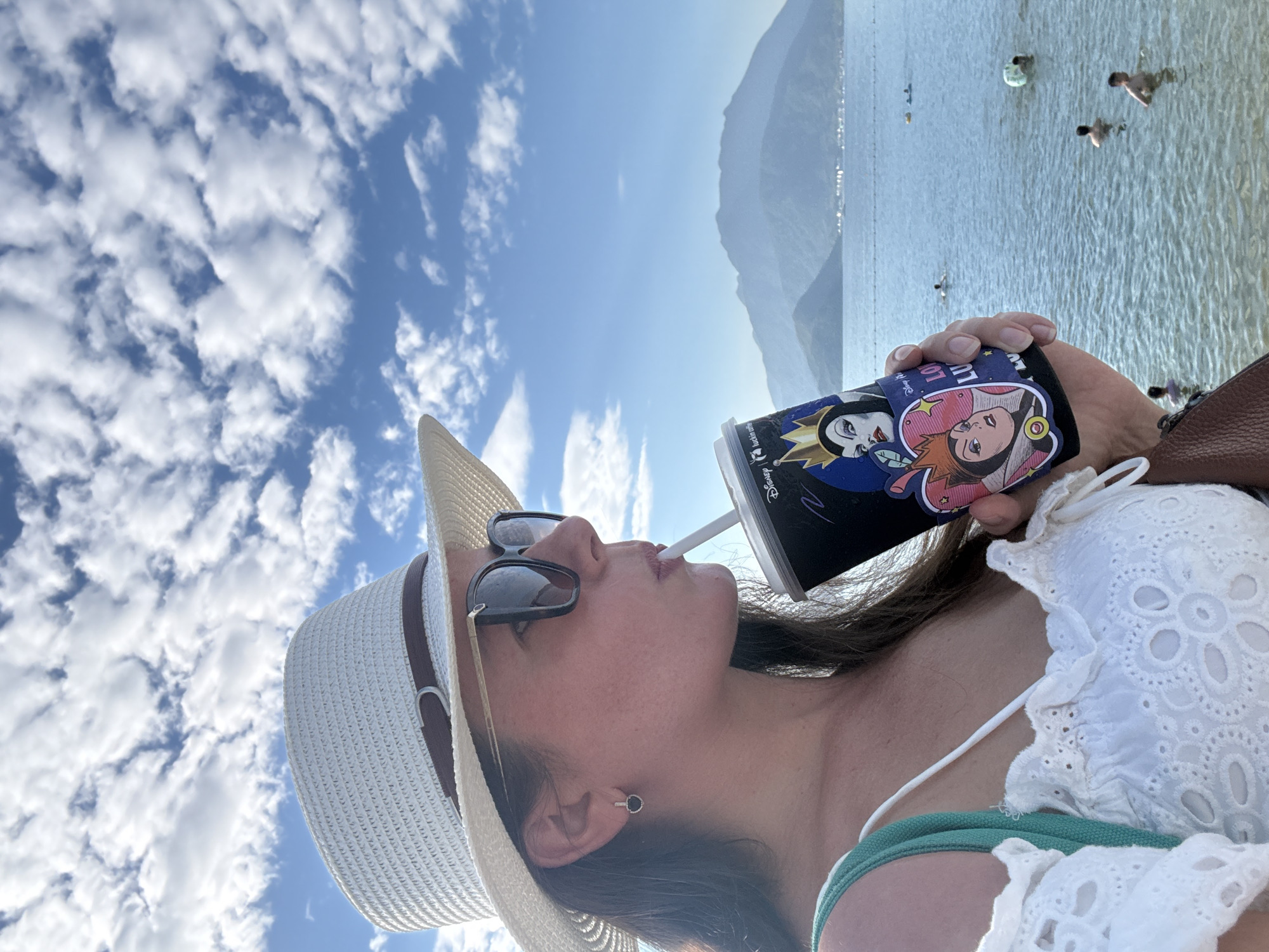 The image shows a woman wearing a white sun hat and sunglasses, drinking from a disposable cup with a straw. She is dressed in a white, off-the-shoulder top with eyelet detailing. The cup features images of Disney villains. She is standing near a body of water, which appears to be a beach or bay. In the background, there are mountains and a blue sky with white clouds. Several people are visible in the water, some swimming and others standing. The scene is likely located in a coastal area within the city of Longgang District, China, given the natural landscape and the presence of other people enjoying recreational activities in the water.