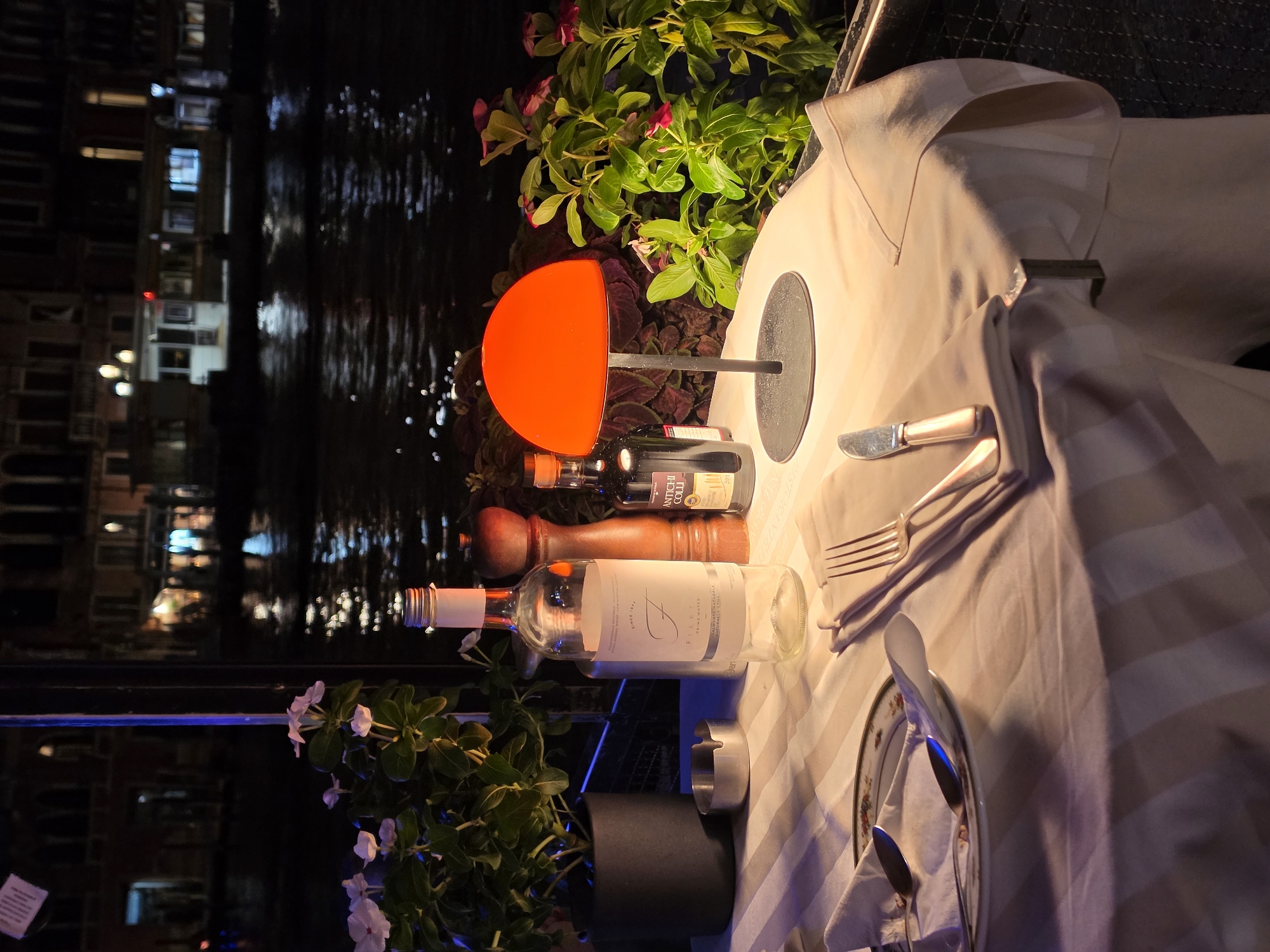 The image depicts an outdoor dining table set for one person at night, positioned alongside a canal in Venice, Italy. The table is covered with a striped tablecloth and features a plate with two spoons, a folded napkin, and a fork and knife. On the table, there's a bottle of water, a wooden pepper grinder, and a small bottle of what appears to be olive oil. An orange-hued table lamp illuminates the setting, casting a warm glow. Behind the table, lush green plants with small pink flowers are visible. The canal water reflects the lights of the buildings across the way, creating a serene and romantic ambiance. The buildings across the canal appear to be historic Venetian structures.