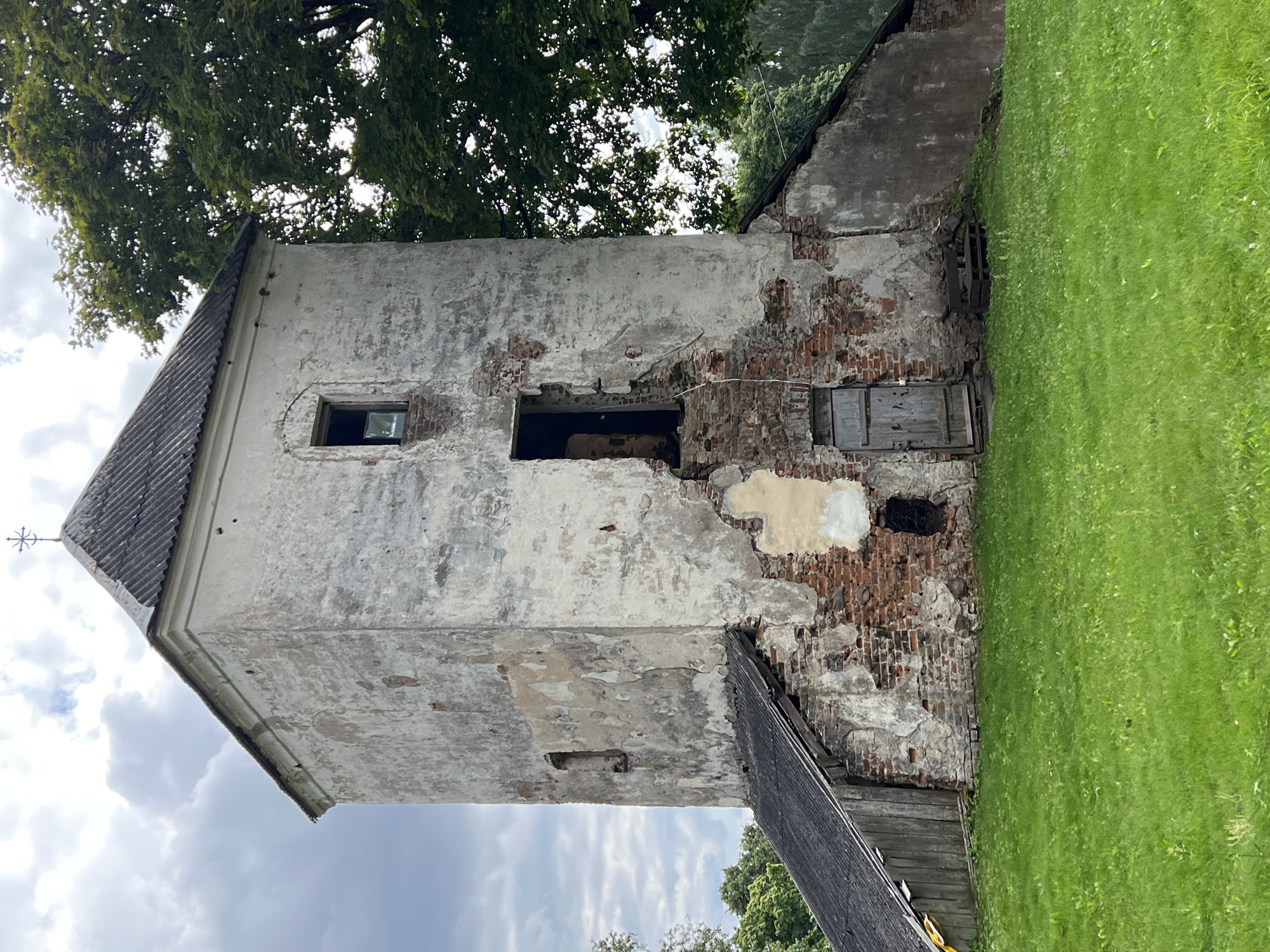 The image shows an exterior view of the Norviliškės Castle, a historical landmark in Norviliškės, Lithuania. The castle appears to be in a state of disrepair, with weathered white stucco peeling off to reveal red brick and stone underneath. The main structure is a tall, square tower with a pitched, grey, corrugated roof topped by a cross. There are several openings in the tower, including a small, rectangular window and a larger, arched doorway that appears to be open. A smaller, wooden door is visible at the base of the tower. The tower is attached to a lower section with a similar dark, corrugated roof that extends to the left and right. A well-maintained green lawn surrounds the base of the structure, and lush green trees are visible in the background, suggesting a rural or park-like setting. The sky is overcast with scattered clouds. There are no people visible in the image.