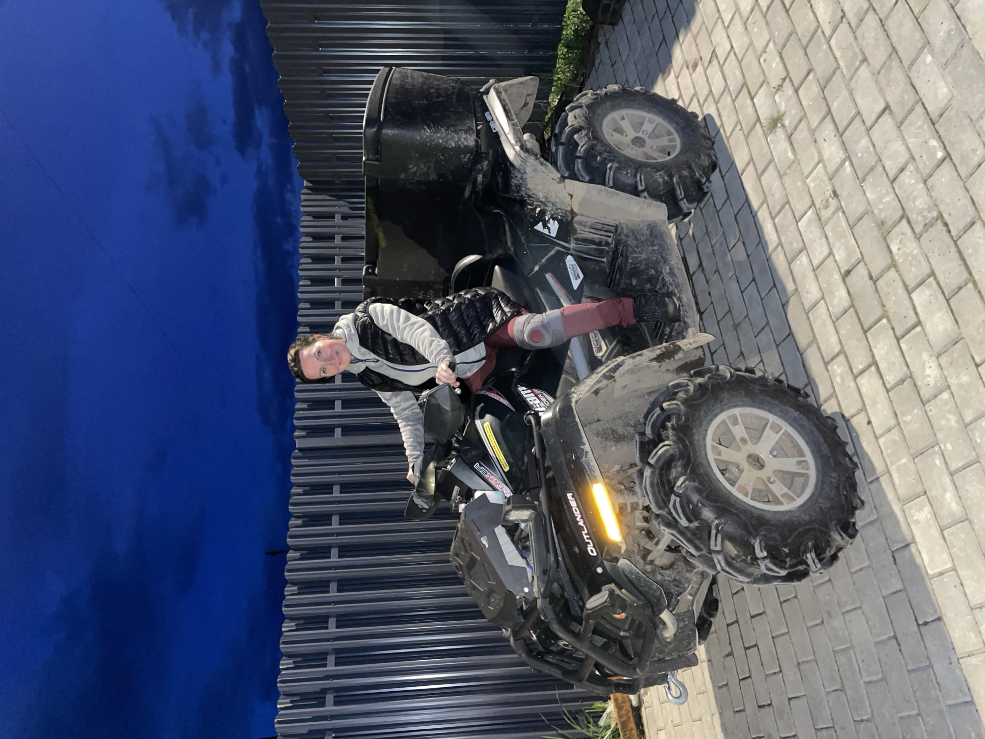 The image shows a woman sitting on a black Can-Am Outlander ATV. She is wearing a gray and white jacket and maroon pants. The ATV is parked on a paved surface made of rectangular blocks, likely a driveway or walkway. A corrugated metal fence is visible in the background under a twilight sky. The location is Solov'evka, Russia.