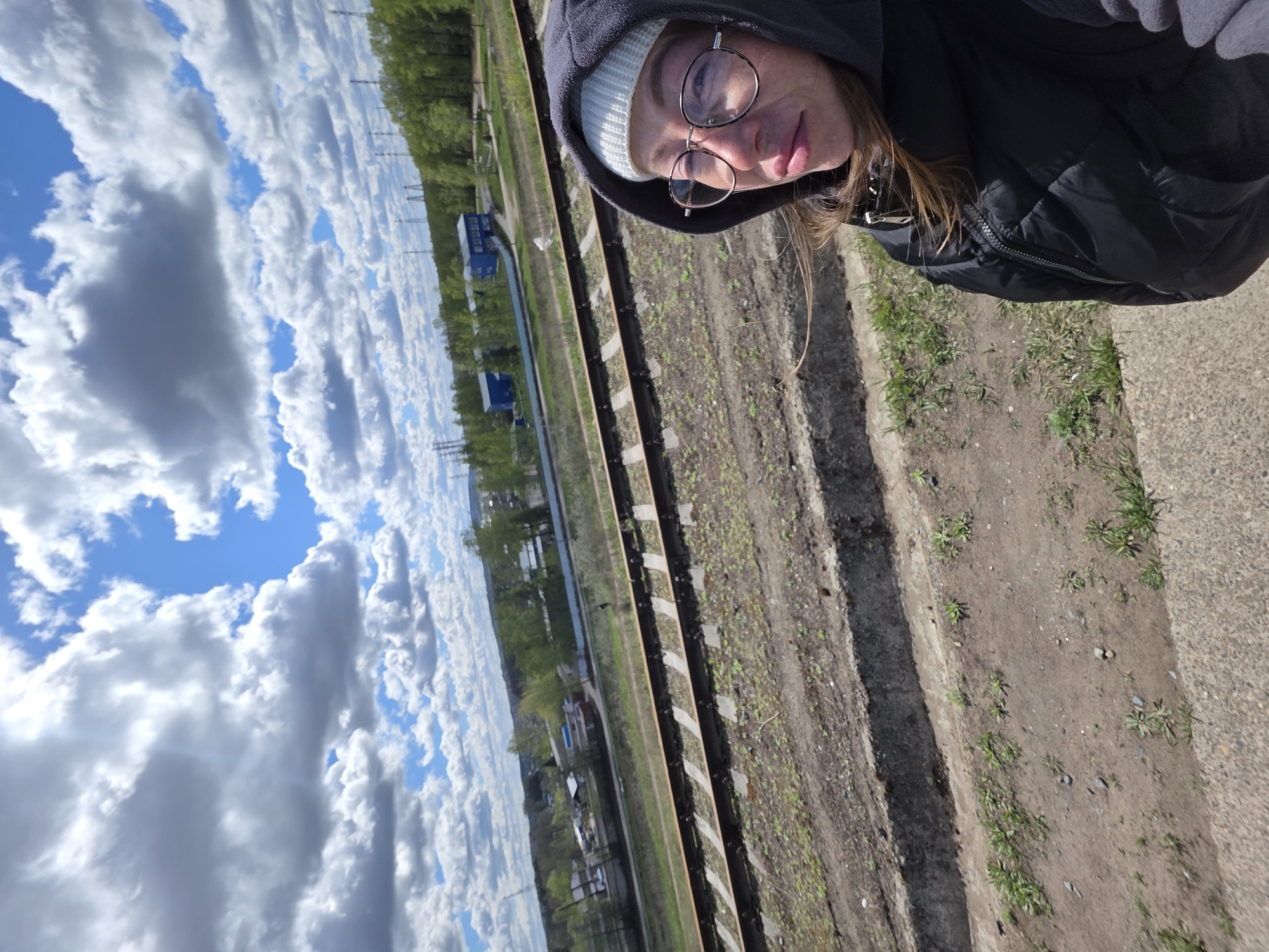 The image is a selfie taken by a woman in a hooded jacket and glasses, standing next to a railway track. She wears a white beanie under the hood. The track runs diagonally across the frame, with vegetation growing between the rails. Beyond the track, there are glimpses of a town or village under a bright sky with puffy white clouds.
Based on the visible elements, including the type of railway infrastructure and the appearance of the town in the background, it is possible the location is near Chaikovsky, Russia. This inference is based on the assumption the image was taken in the context described in the prompt.