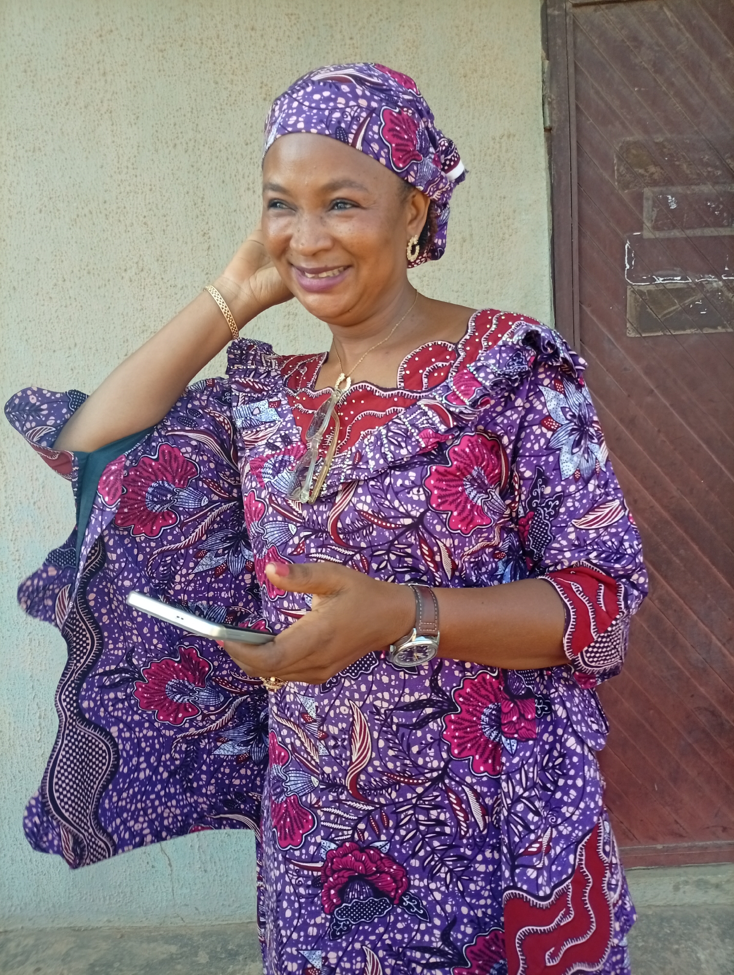 A medium shot captures a Black woman in her late 40s or 50s, wearing a vibrant purple and pink floral patterned traditional African dress and headwrap. She is standing outside a building, with a textured light beige wall behind her to the left and a dark brown wooden door to her right. The woman is smiling and looking to her left, with her right hand resting behind her head, and her left hand holding a silver smartphone. She is wearing a gold bracelet on her left wrist and a silver watch on her right wrist. A gold necklace with a pendant hangs around her neck. The lighting suggests it is daytime, possibly outdoors. The image appears to be taken in the Sabon Tasha area of Kaduna State, Nigeria, given the architectural style and the woman's attire, suggesting a residential or community setting within the city.