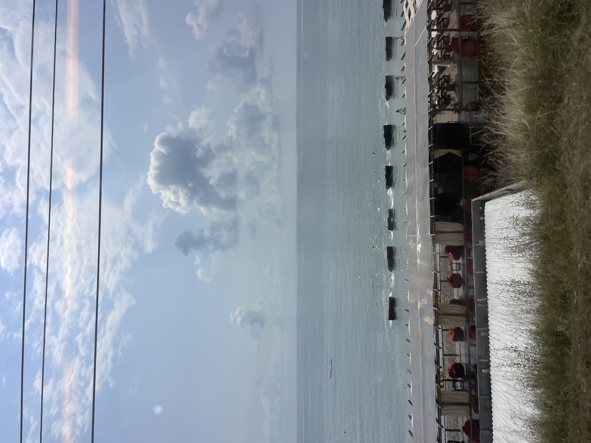 The image depicts a seaside scene, likely in Sochi, Russia, featuring a beachside establishment against a backdrop of the sea and sky. In the foreground, dry grass and some vegetation are visible. Beyond this, a white, corrugated roof structure is partially seen. Adjacent to it is a wooden deck covered by a translucent roof, supported by wooden posts. This area is furnished with red beanbag chairs and appears to be a seating or lounging area for a cafe or restaurant. The beach itself is rocky, with dark, submerged structures in the water that may be part of a breakwater or jetty. The sea is calm and a light blue, with a few small waves breaking near the shore. In the distance, two small boats are visible on the water. The sky is a light blue, dotted with fluffy white clouds. Several parallel black wires stretch across the upper portion of the frame, suggesting the photo was taken from a moving vehicle or a location with overhead power lines.