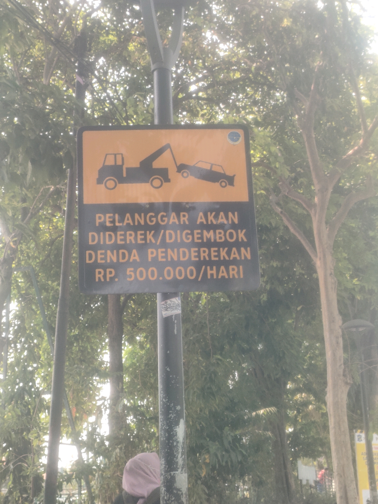 A street sign is displayed on a metal pole, featuring a yellow rectangular top with a black illustration of a tow truck towing a car. Below this, a black rectangular section contains text in yellow lettering. The text reads: "PELANGGAR AKAN DIDEREK/DIGEMBOK DENDA PENDEREKAN RP. 500.000/HARI". This translates to "Violators will be towed/locked, towing fine RP. 500,000/day". The sign is situated outdoors, with lush green trees and foliage forming the background. The scene appears to be during daylight, with sunlight filtering through the leaves, creating a somewhat hazy effect. The sign is located in the city of Surabaya, Indonesia.